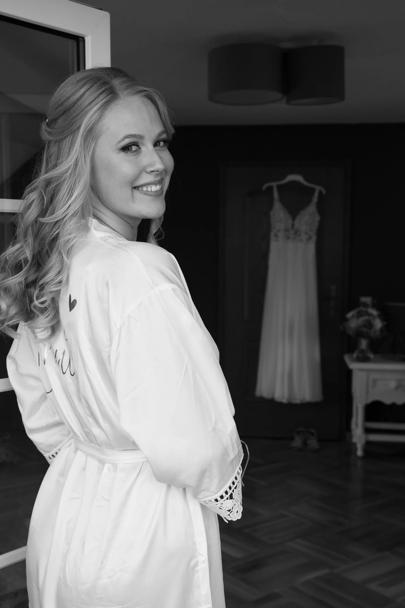 Schöne junge Frau in einem weißen Satin-Badrock, lächelnd, im Hintergrund ein Hochzeitskleid auf einem Kleiderbügel in einem dunklen Raum.