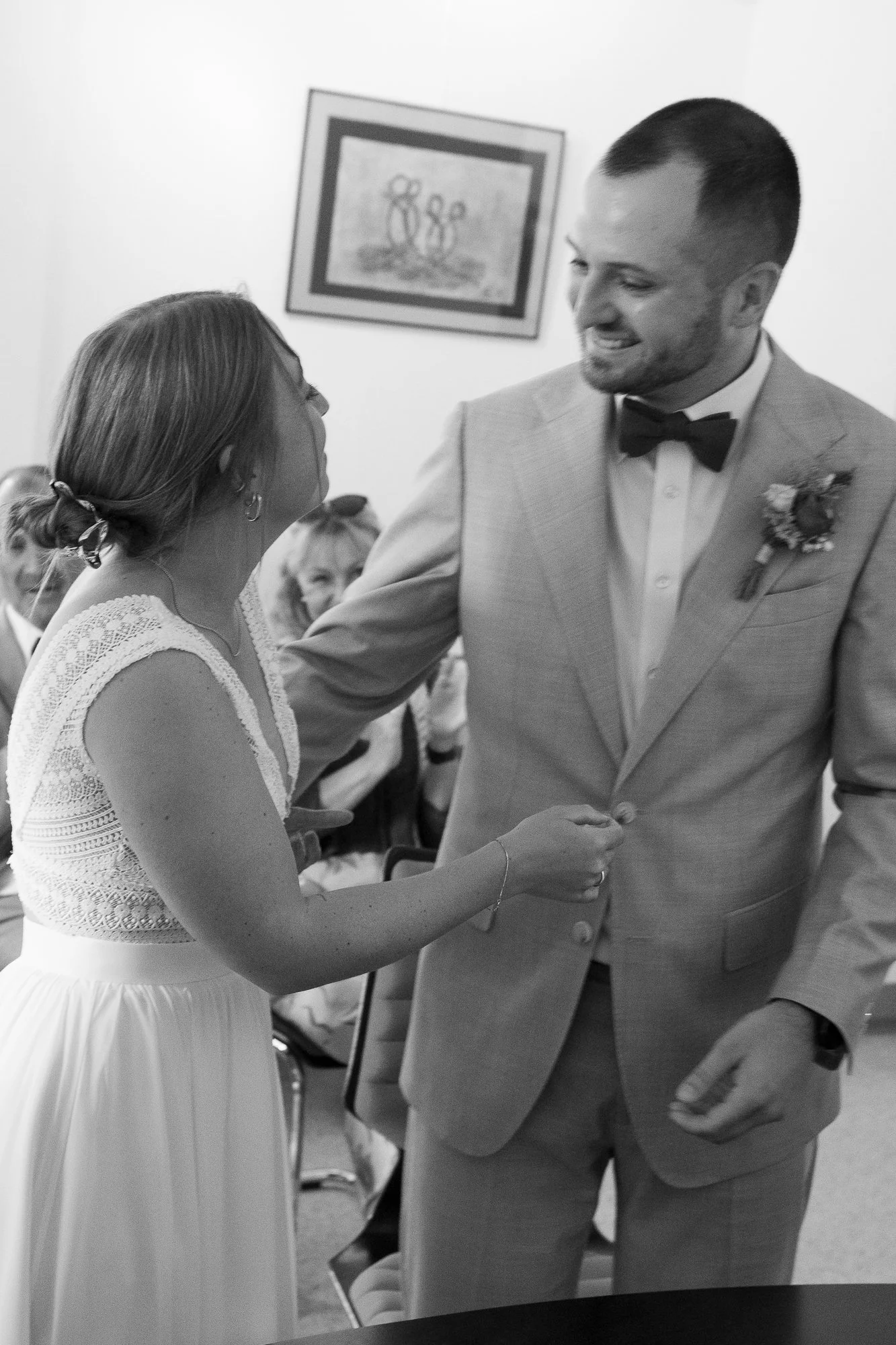 Ein Brautpaar bei der Hochzeit, der Bräutigam trägt einen Anzug mit Fliege, die Braut trägt ein weißes Kleid, sie halten Händchen und lächeln sich an, im Hintergrund sitzen Hochzeitsgäste, die Wand hat ein Bild.