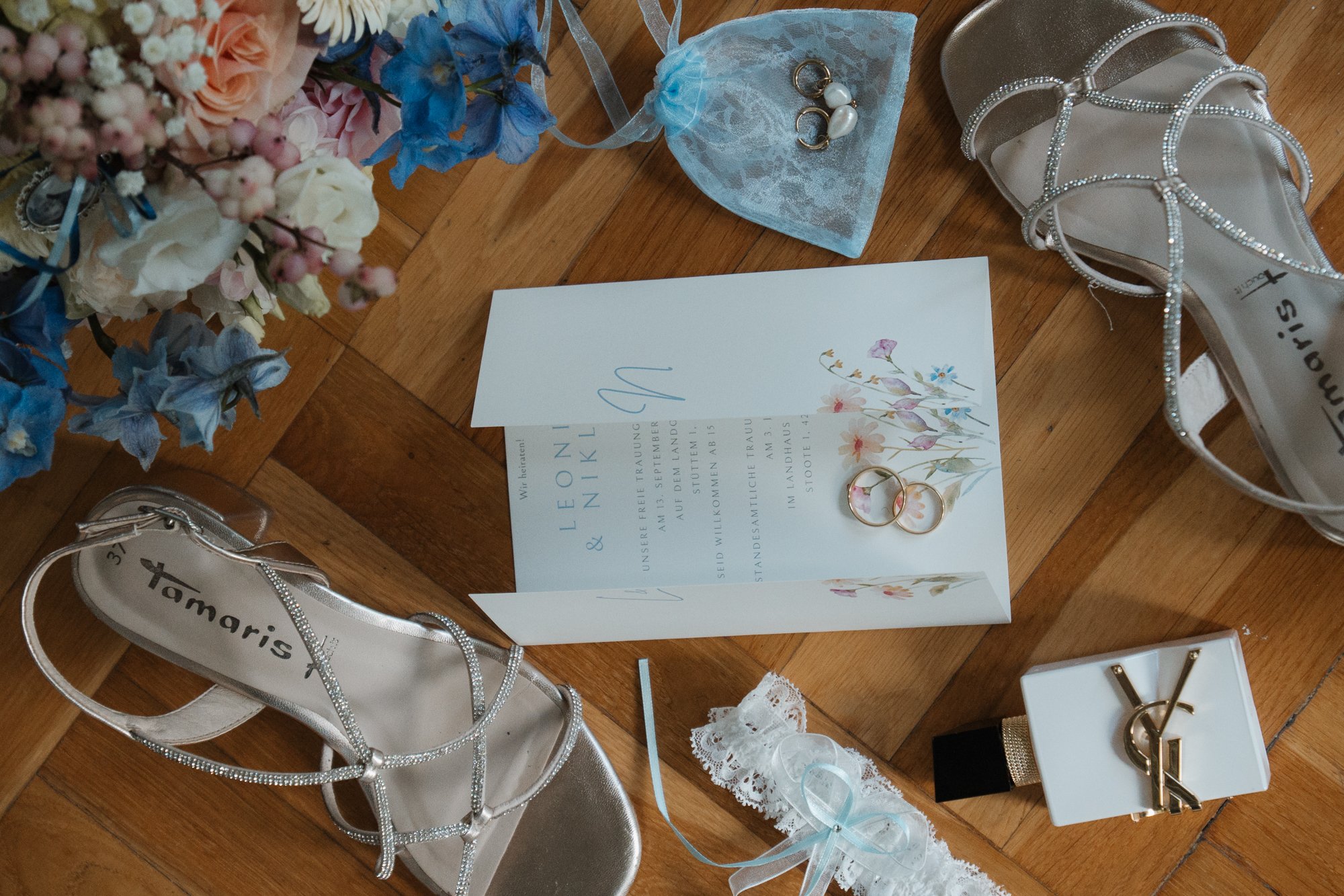 Hochzeitsaccessoires einschließlich silberne High Heels mit Perlenapplikationen, Hochzeitskarte mit Blumenmuster, Trauringe, Brautschleier mit blauer Schleife, Blumenteile, Parfümflasche, Schmuckstück, und Brautstrauß auf Holztisch.