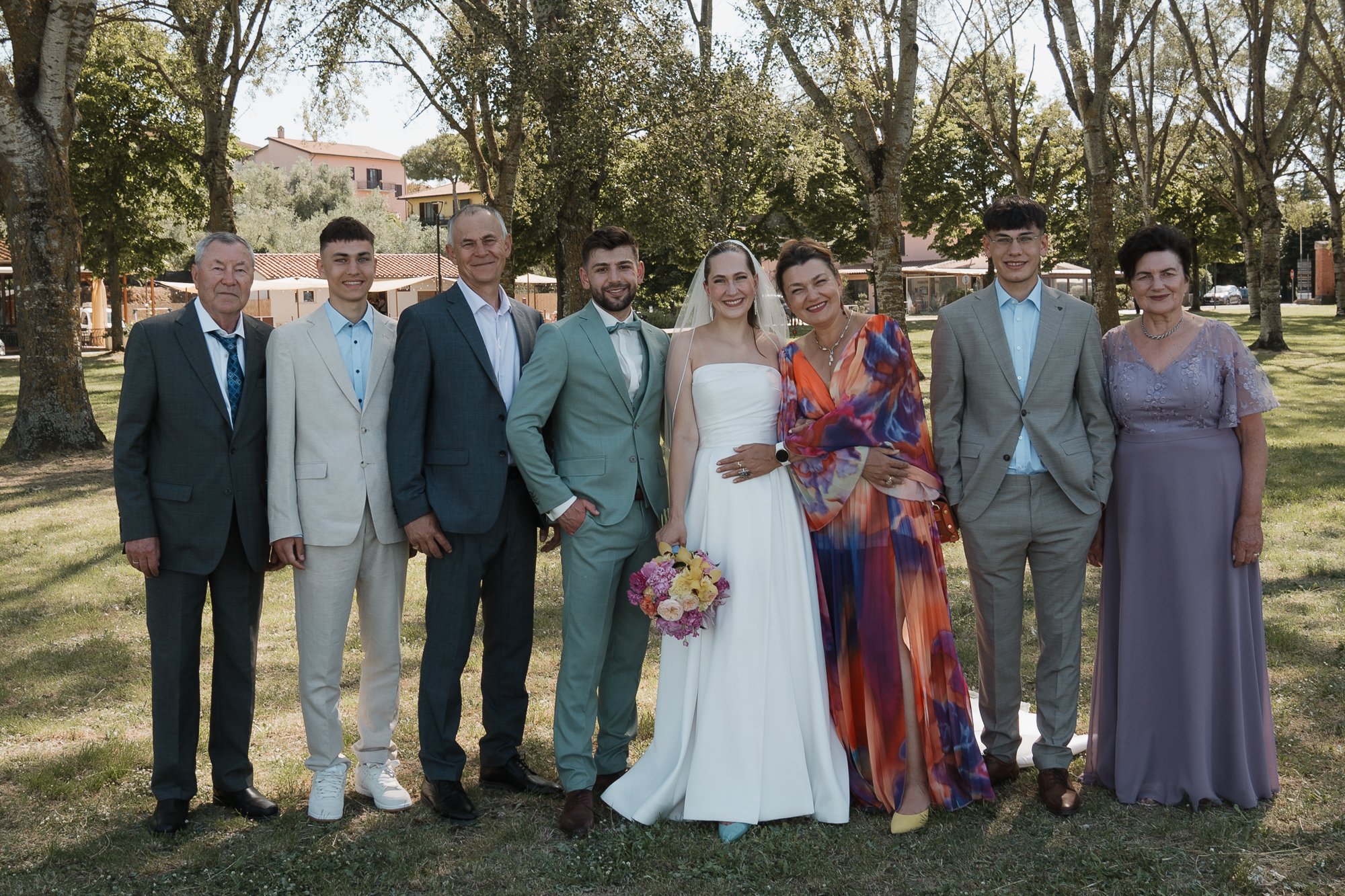 Gruppe von acht Menschen bei einer Hochzeitsfeier im Freien, inklusive Braut in Weiß mit Blumenstrauß, Bräutigam in Anzug, und weitere Gäste in formeller Kleidung, vor Bäumen und Gebäuden im Hintergrund.