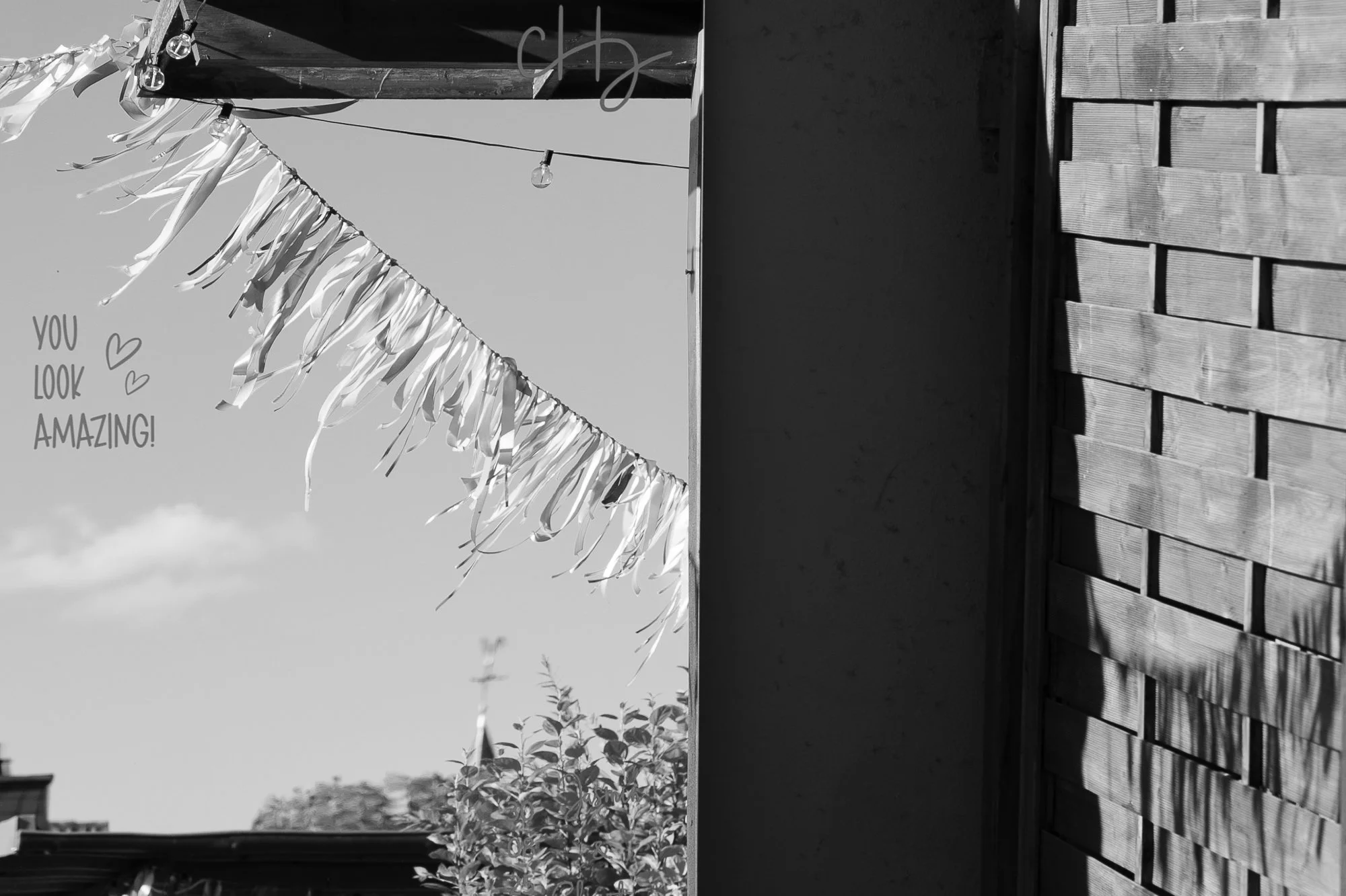 Schwarz-weiß-Foto eines bemalten Wandabschnitts mit einem Spruch: "You look amazing!" und kleinen Herzen. Im Hintergrund sind eine Leiter, eine Fensterscheibe mit Glühbirne, Pflanzen und Häuser zu sehen.