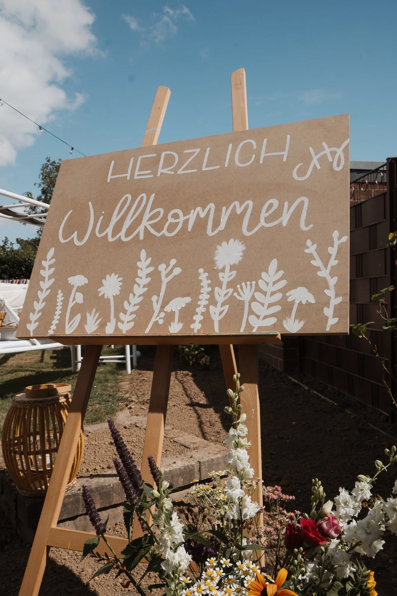 Ein Willkommensschild mit dem deutschen Text "Herzlich Willkommen" und weißen Blumenmotiven, aufgestellt vor einem Garten mit Blumen und einem blauen Himmel.