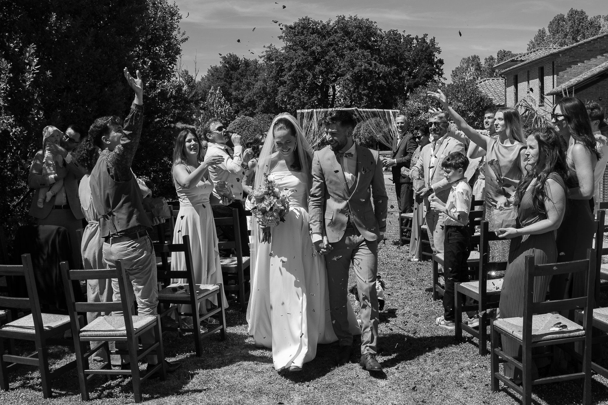 Ein Hochzeitsbild in Schwarz-Weiß zeigt ein Brautpaar, das Hand in Hand geht, umgeben von fröhlichen Gästen im Freien bei Sonnenschein, mit einigen Gästen, die Konfetti werfen.