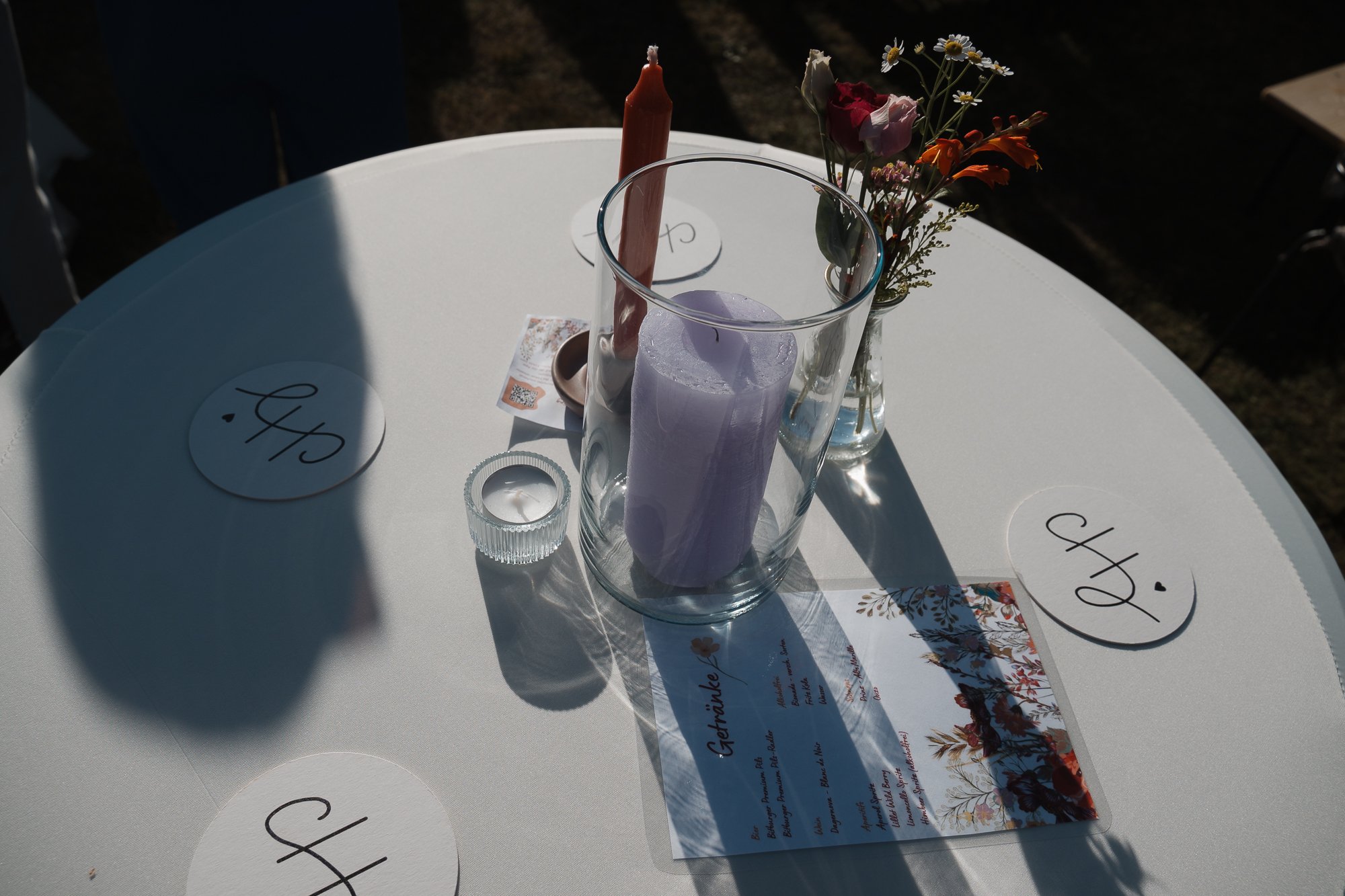 Ein runder weißen Tisch mit Kerzen, Blumen in einer Vase, und Platzkarten. Es gibt eine rote Kerze, zwei lilafarbene Kerzen, eine kleine weiße Kerze, sowie menüartige Papiere mit Blumenmuster, die wahrscheinlich eine Menükarte sind. Die Stimmung ist 