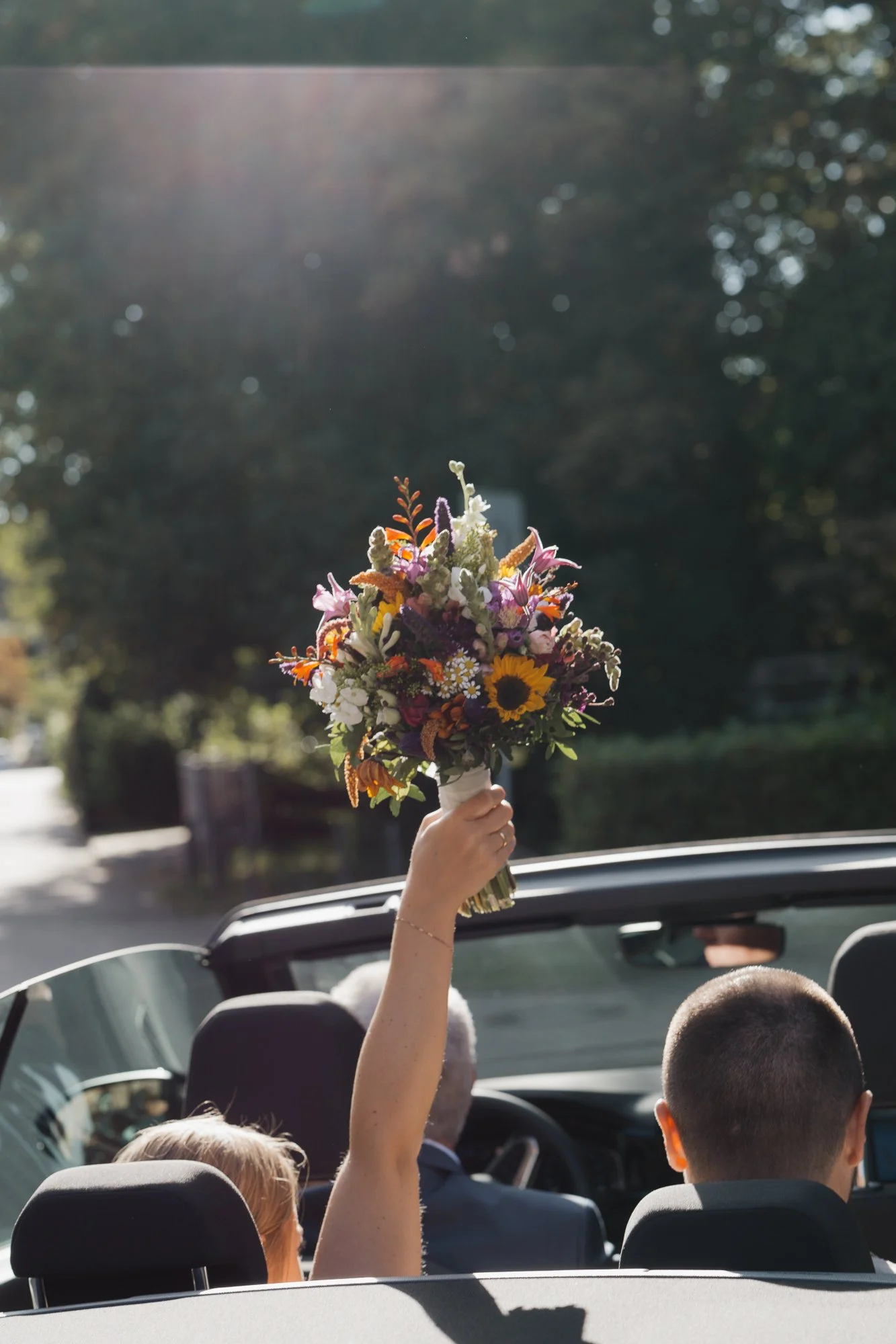 Eine Hand hält einen bunten Blumenstrauß in einem offenen Cabrio während eines sonnigen Tages, im Hintergrund Bäume und Straße.