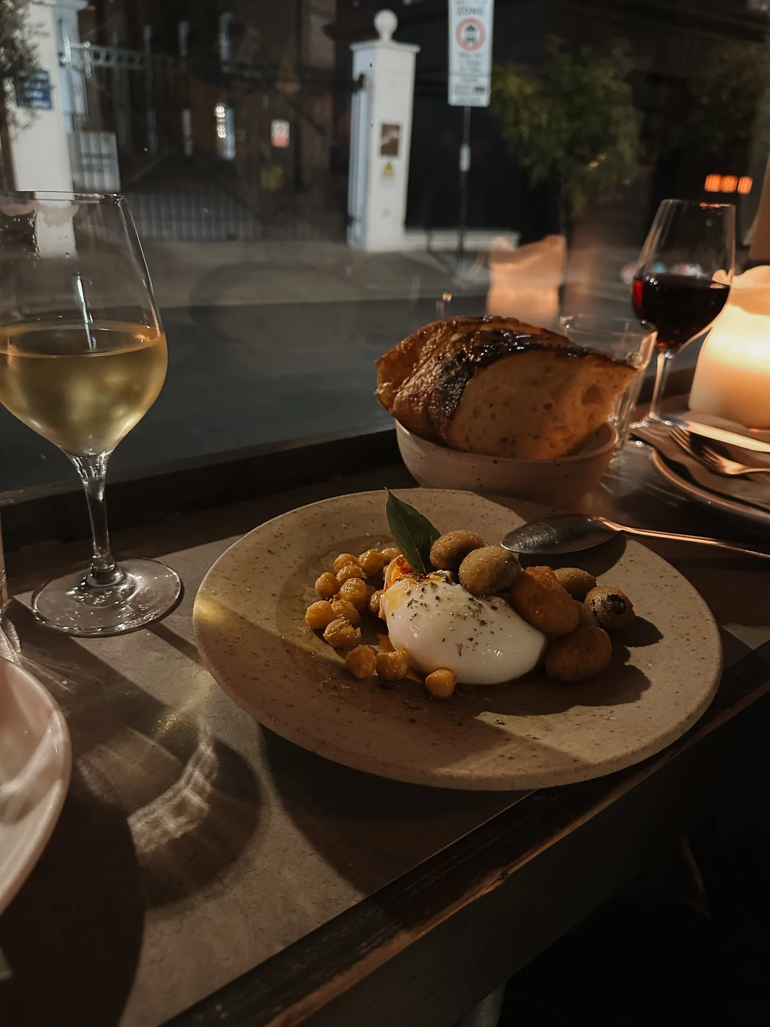 Ein gedeckter Tisch mit einem Teller mit Burrata-Käse, Kichererbsen und Oliven, ein Glas weißer Wein, ein Glas Rotwein, Brot in einer Schüssel, ein brennendes Kerze, im Hintergrund eine Straße bei Nacht.