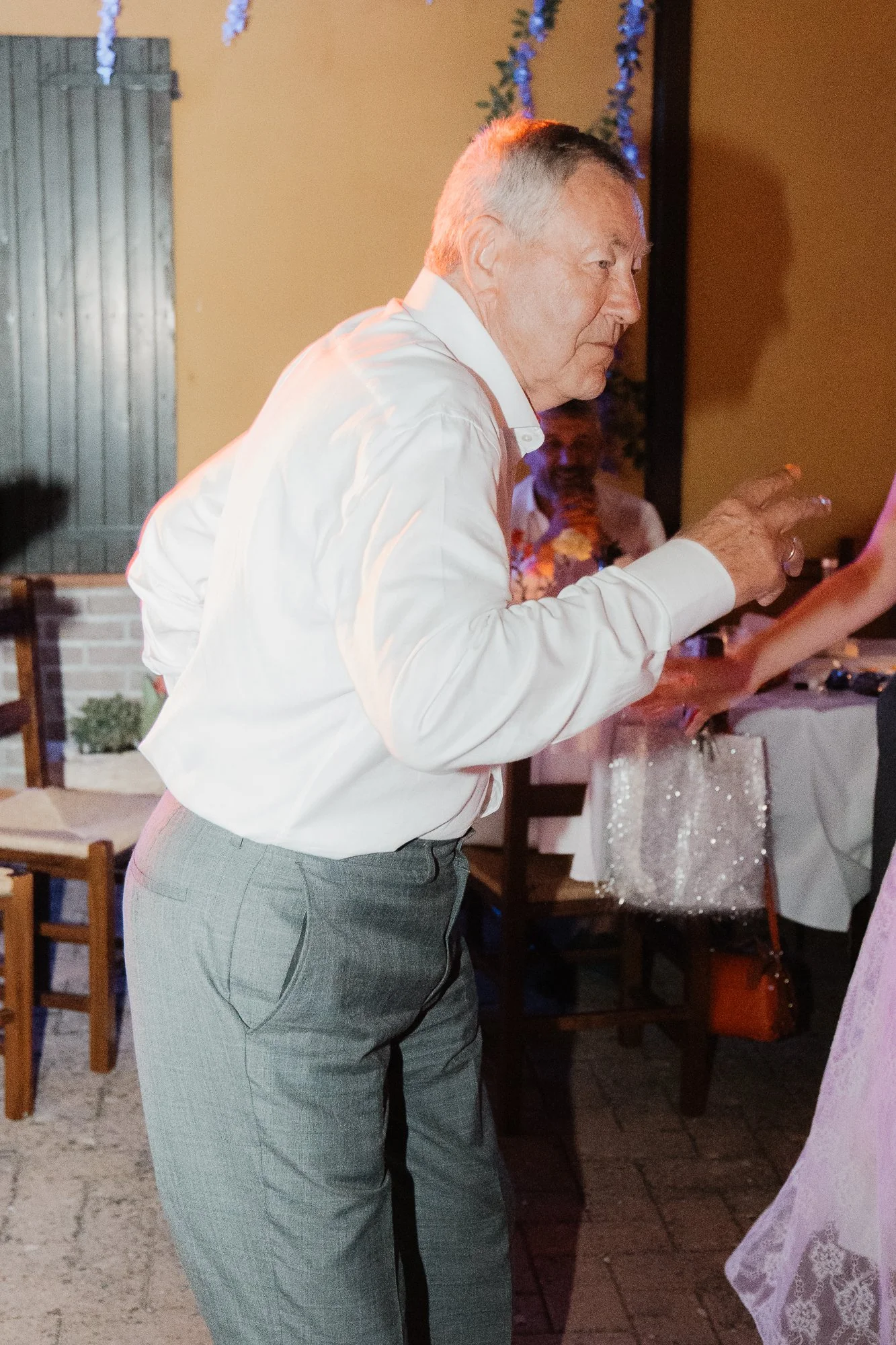 Ein älterer Mann in formeller Kleidung, der auf einer Feier tanzt oder sich bewegt, mit anderen Gästen im Hintergrund in einem festlichen Ambiente.