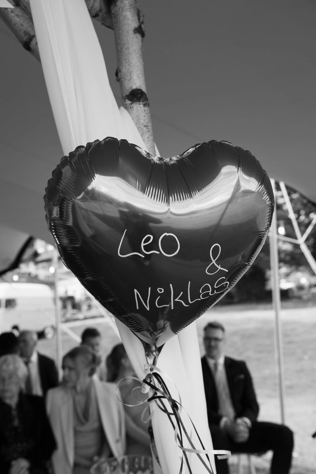 Ein Herzballon mit der Aufschrift 'Leo & Niklas' vor einer Gruppe von Menschen bei einer Feier im Freien, im Hintergrund Bäume und eine weiße Zeltstruktur.