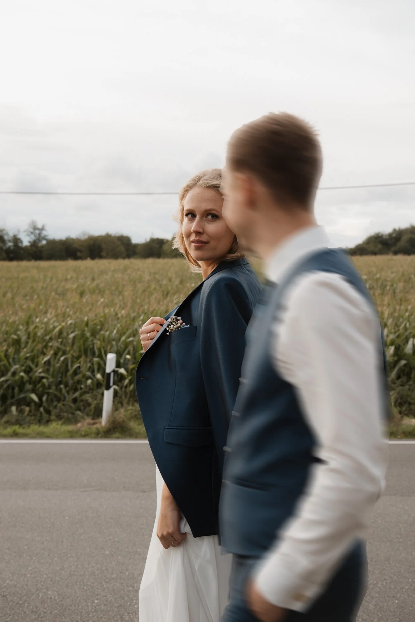 Ein Mann und eine Frau gehen an einer Landstraße entlang, die Frau schaut zurück, während der Mann vorbeigeht, im Hintergrund Felder und Bäume, bewölkter Himmel.