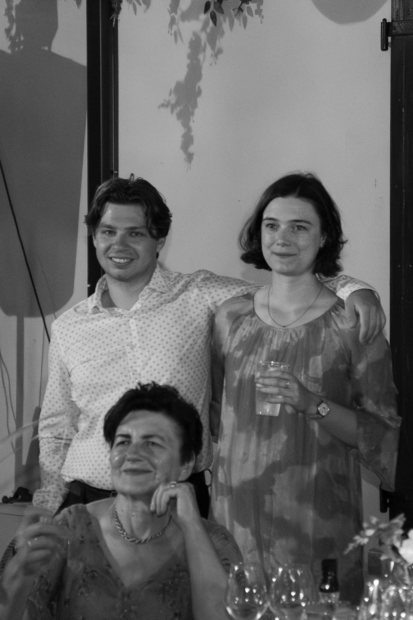 Drei Menschen, zwei Frauen und ein Mann, bei einer Feier, zwei stehen und eine sitzt. Der Mann und die Frau, die steht, haben die Arme um die Schultern des jeweils anderen. Die sitzende Frau hält ein Glas, alle schauen in die Kamera.