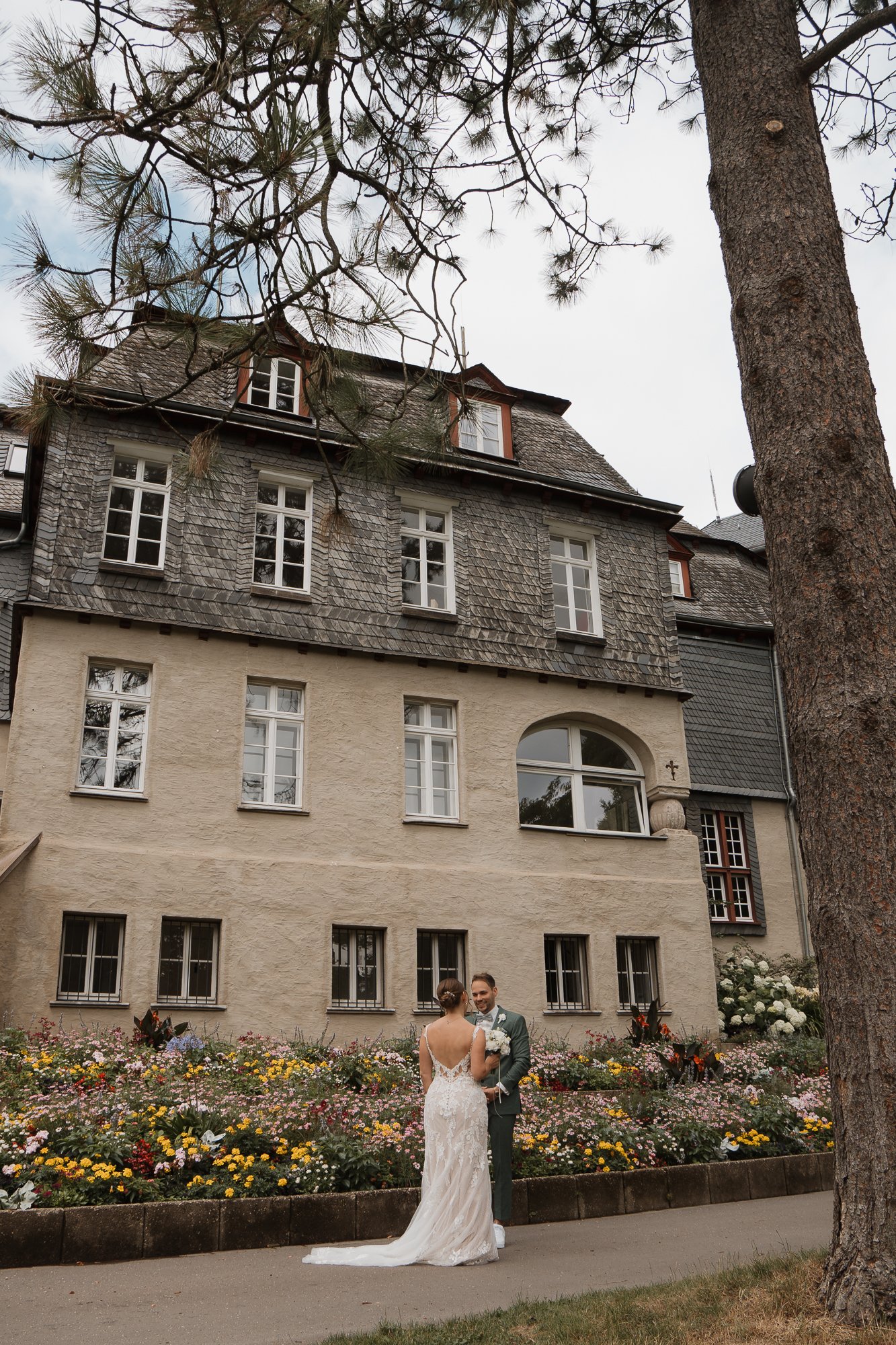 Ein Brautpaar steht vor einem alten Haus mit einer bunten Blumenrabatte im Vordergrund. Das Haus hat zwei bis fünf Stockwerke mit großen Fenstern und einem Schindeldach.