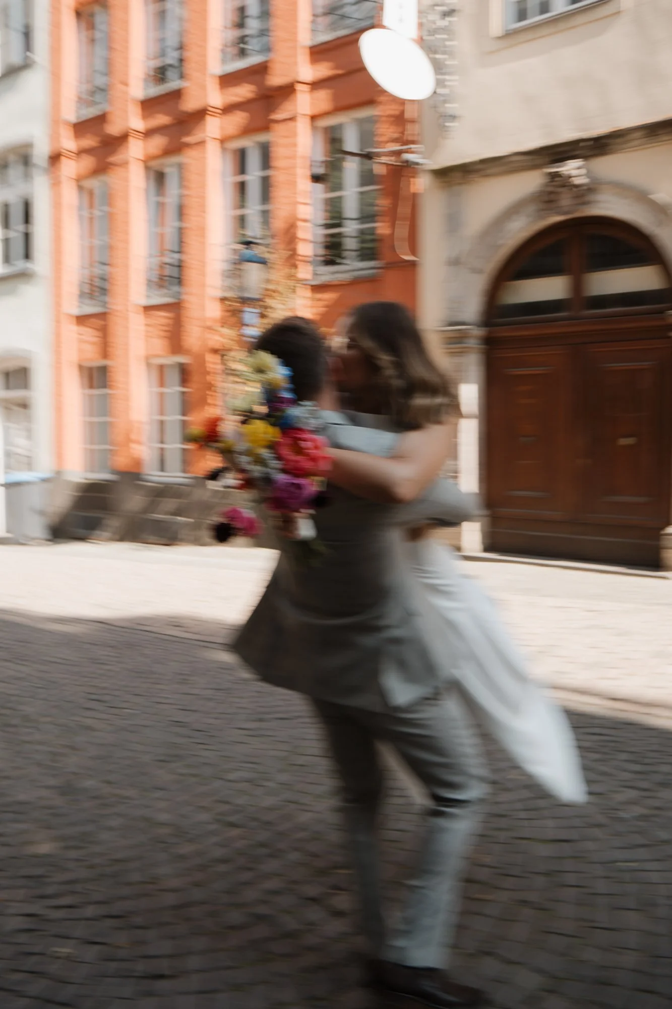 Zwei Personen, die sich umarmen, eine hält einen Blumenstrauß, auf einem Kopfsteinpflaster in einer Stadt mit roten und beigen Gebäuden im Hintergrund.