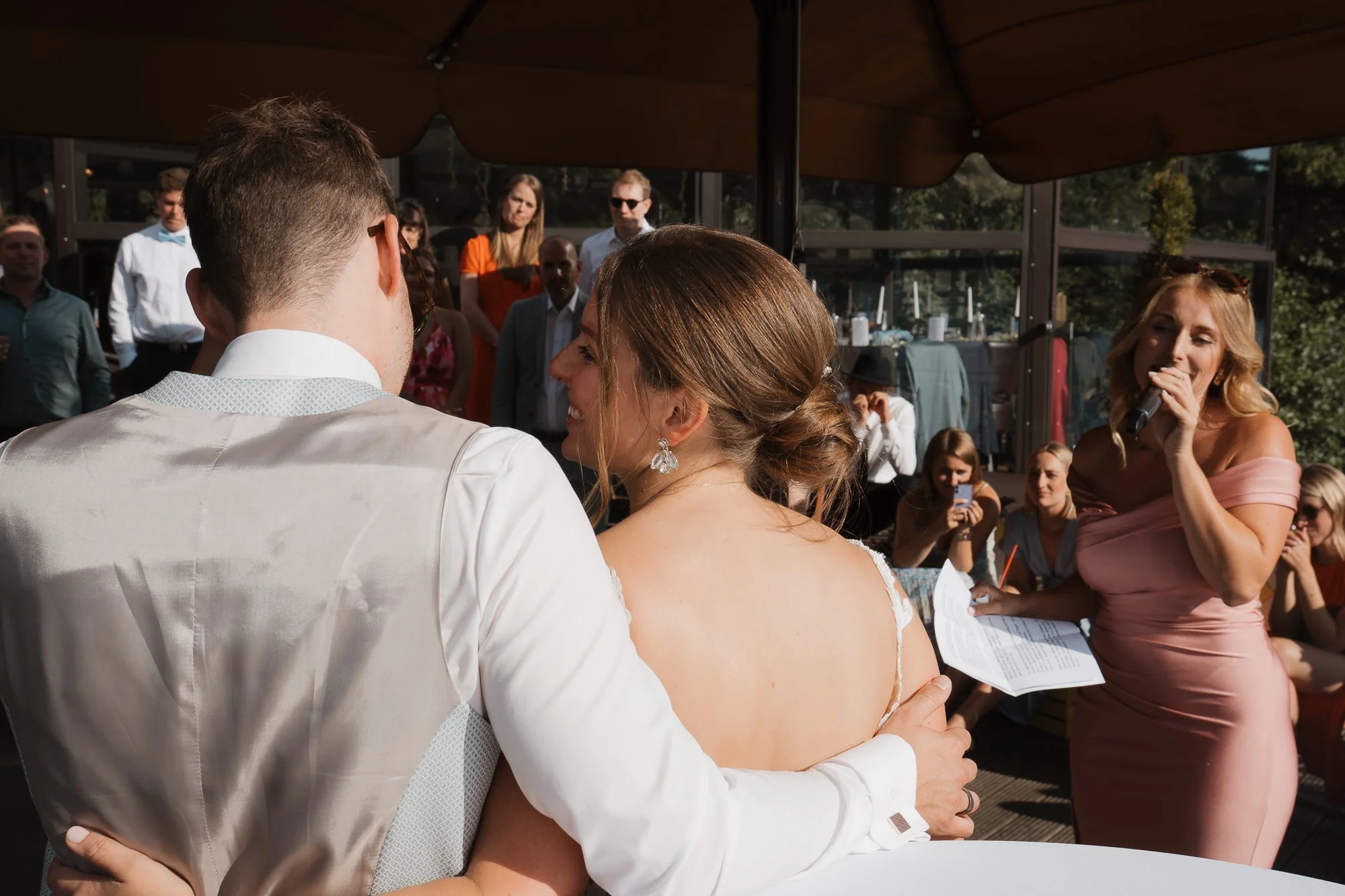 Ein Brautpaar während einer Hochzeit im Freien, umgeben von Gästen, die zuschauen. Die Braut trägt ein weißes Hochzeitskleid und Ohrringe, der Bräutigam in einem hellen Anzug. Eine Frau rechts, vermutlich die Trauungsperson, spricht in ein Mikrofon.