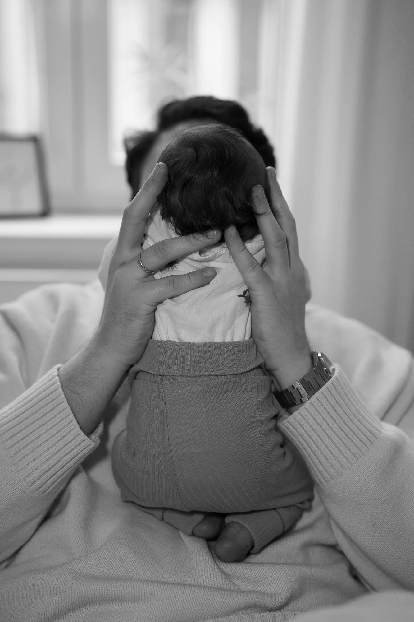 Eine Person hält ein Baby mit dunklem Haar vor dem Gesicht in den Händen, die auf dem Kopf des Babys ruhen. Das Bild ist in Schwarz-Weiß.