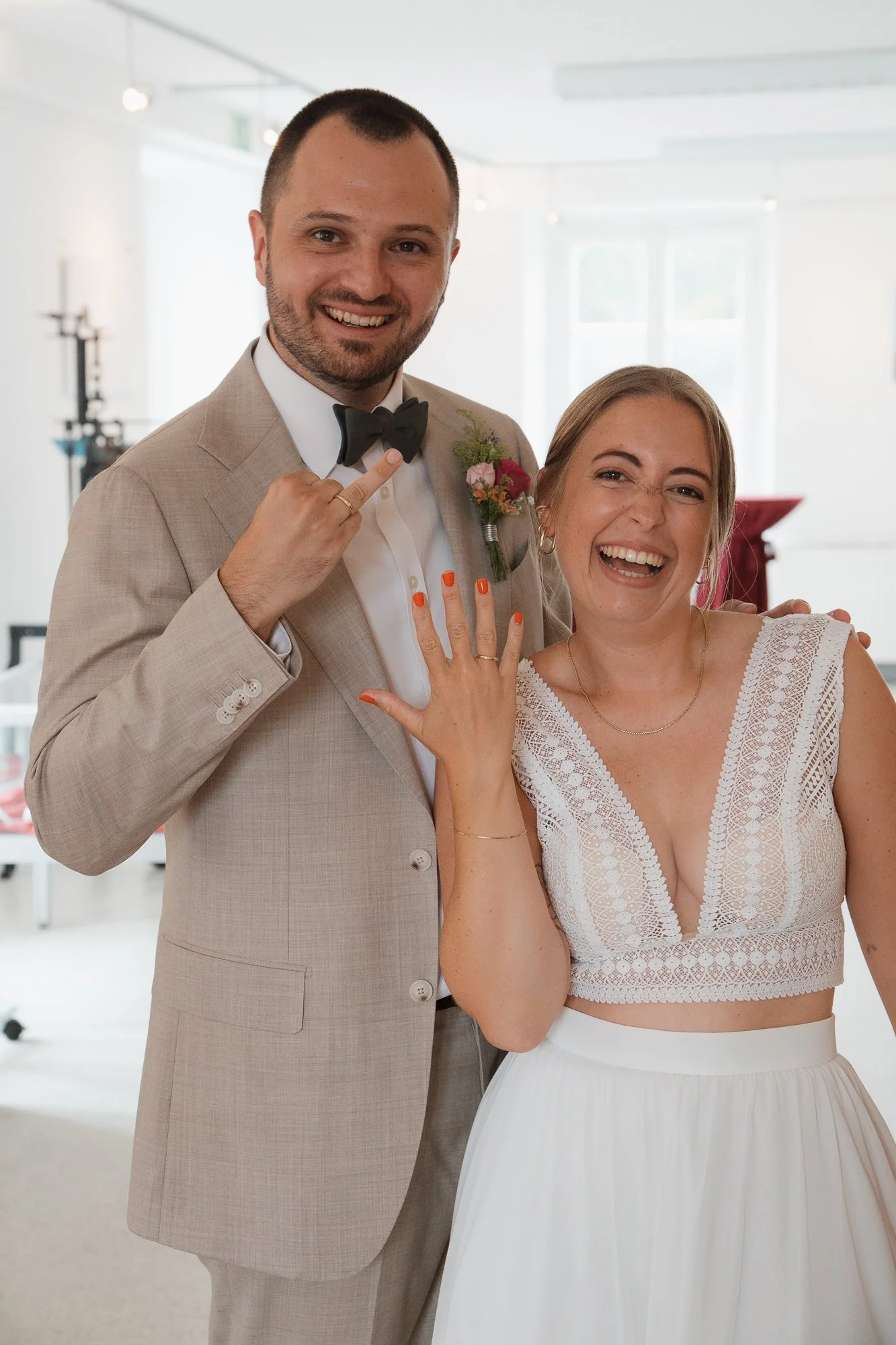 Ein lachendes Paar bei einer Hochzeit, der Mann trägt einen beige Anzug mit Fliege, das Mädchen trägt ein weißes Brautkleid mit Spitze. Sie zeigen ihre Eheringe und machen ein Zeichen mit der Hand.