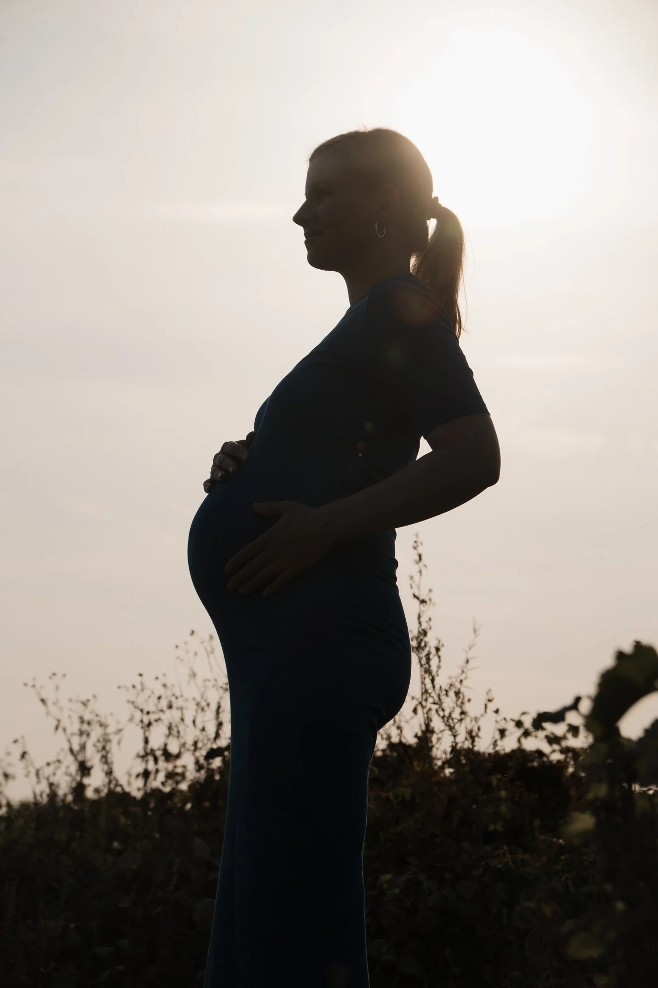 Silhouette einer schwangeren Frau im Freien bei Sonnenuntergang, mit Pflanzen im Hintergrund.
