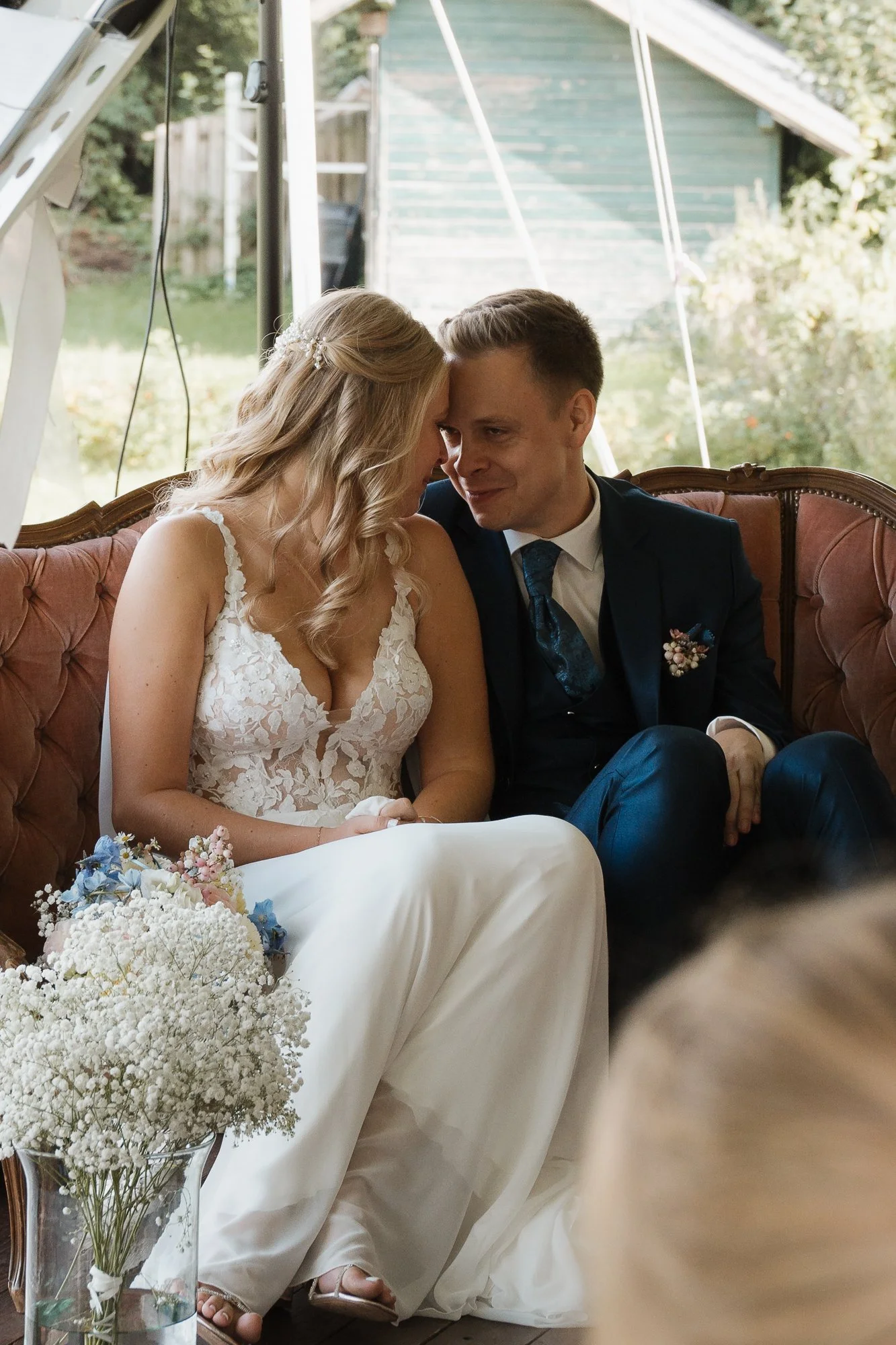 Ein Brautpaar bei ihrer Hochzeit, sitzend auf einer Couch, vergeigen sich aneinander und lächeln.