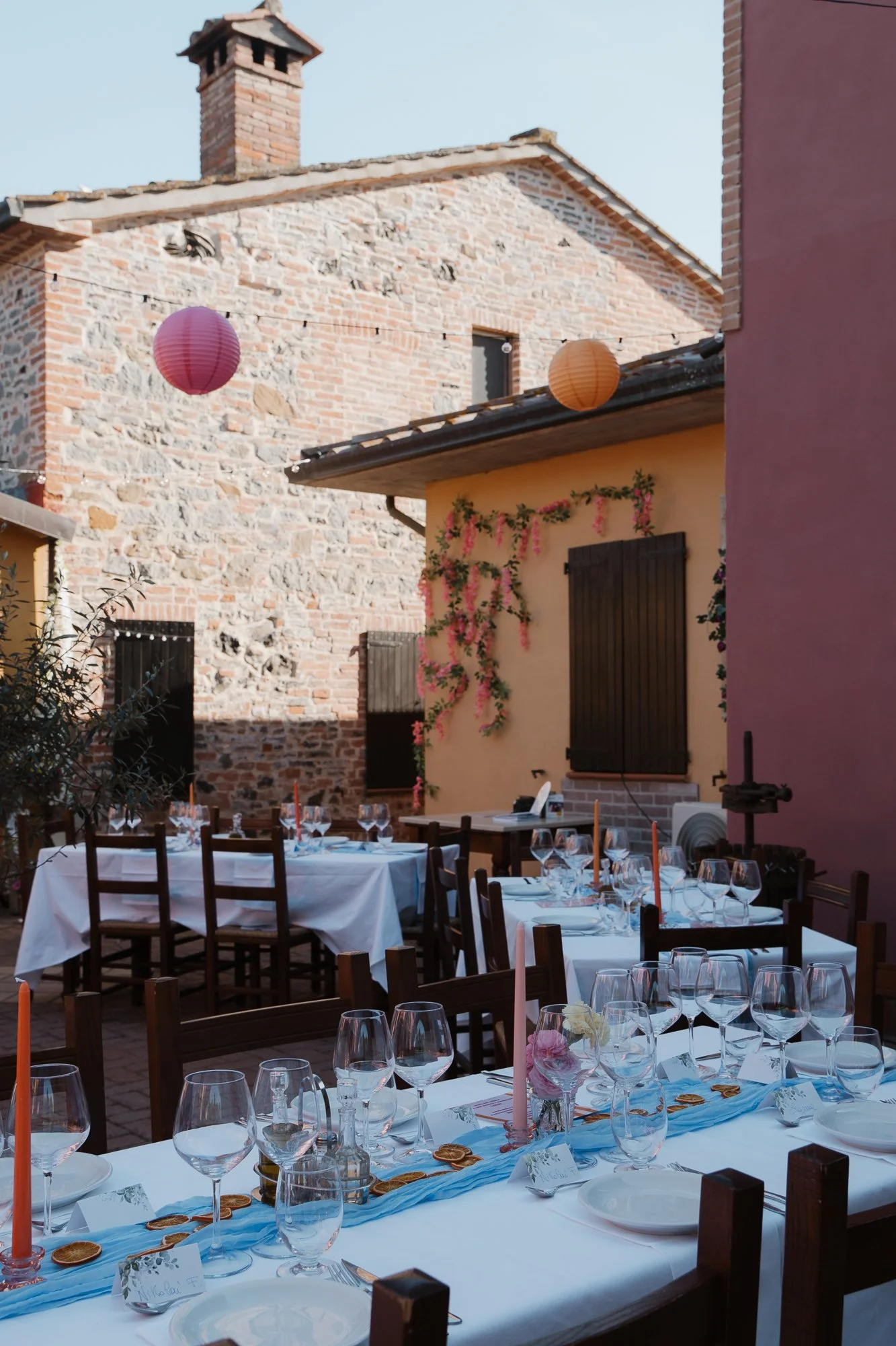 Ein im Freien gedeckter Tisch für eine Feier mit weißer Tischdecke, Kerzen, Gläsern, Geschirr und Dekorationen, vor einem Haus mit bunten Wänden und Gartendekorationen.
