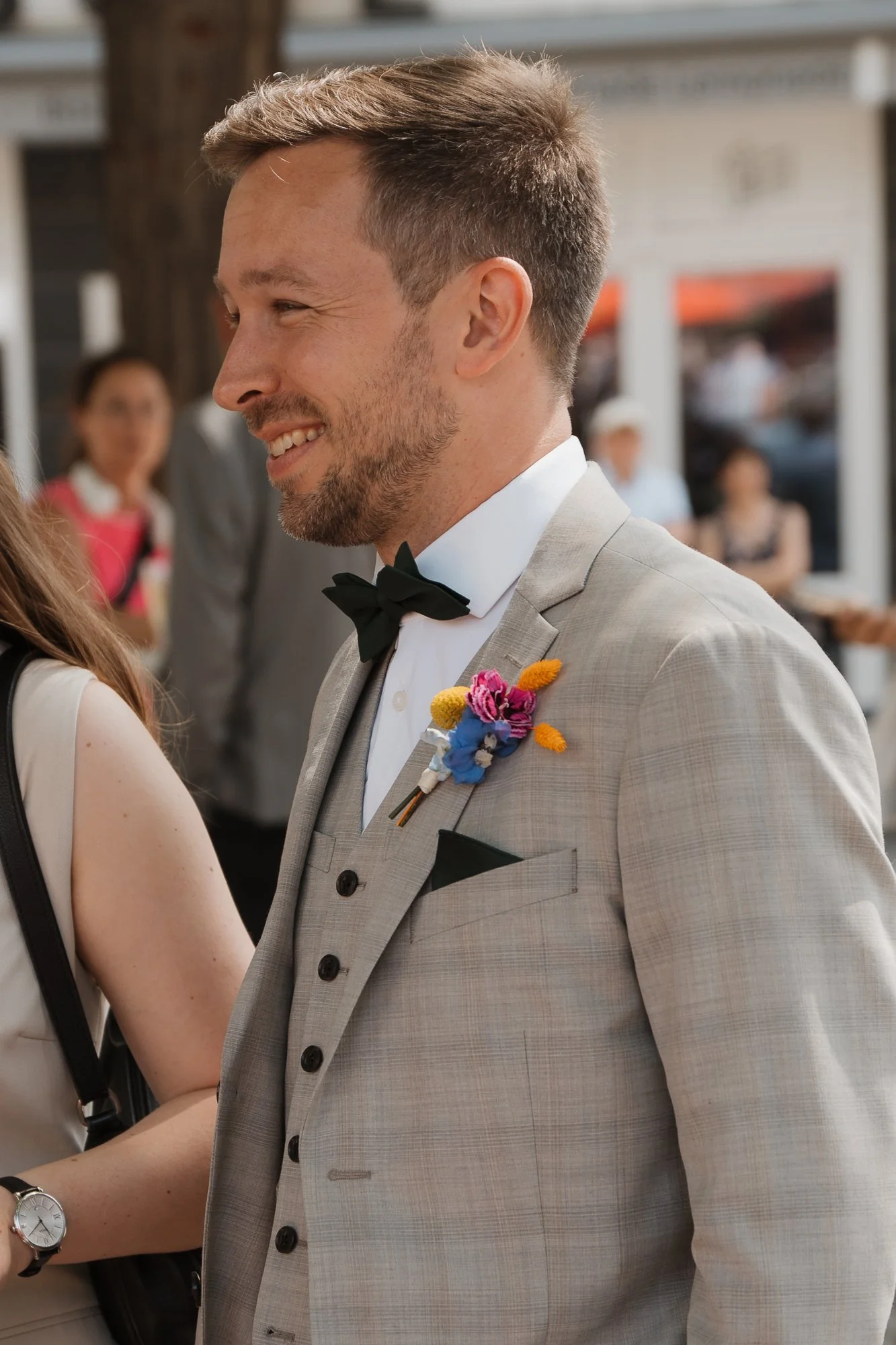 Ein lachender Mann in einem hellegrauen Anzug mit schwarzer Fliege trägt eine bunten Blumenanstecknadel auf der Brust. Im Hintergrund sind andere Personen bei einer Veranstaltung im Freien zu sehen.