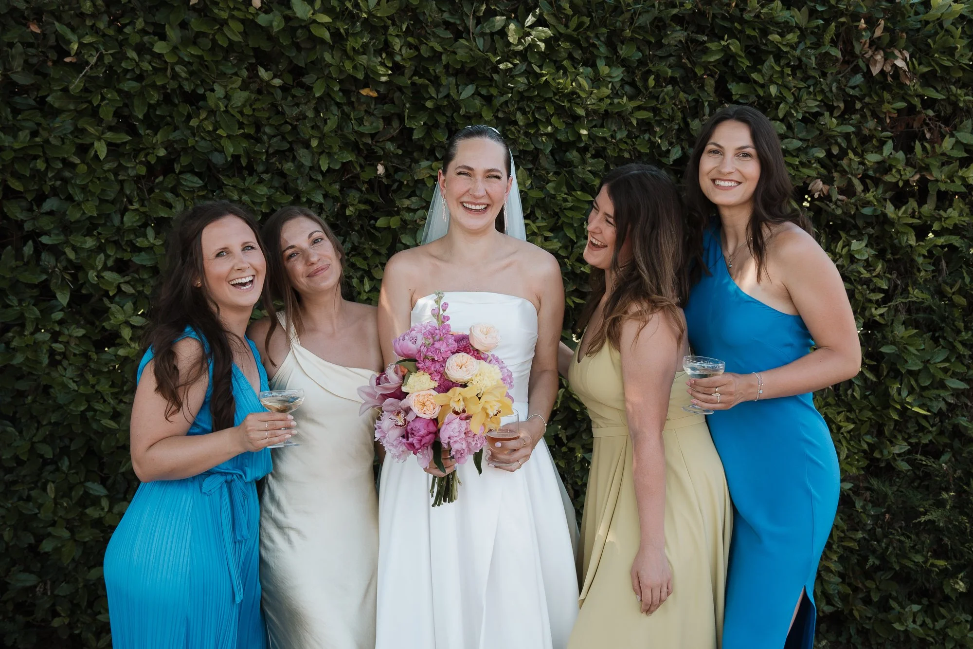 Fünf Frauen bei einer Hochzeit, eine in einem weißen Brautkleid mit Blumenstrauß, die anderen in bunten Kleidern, vor einem grünen Laubwand.