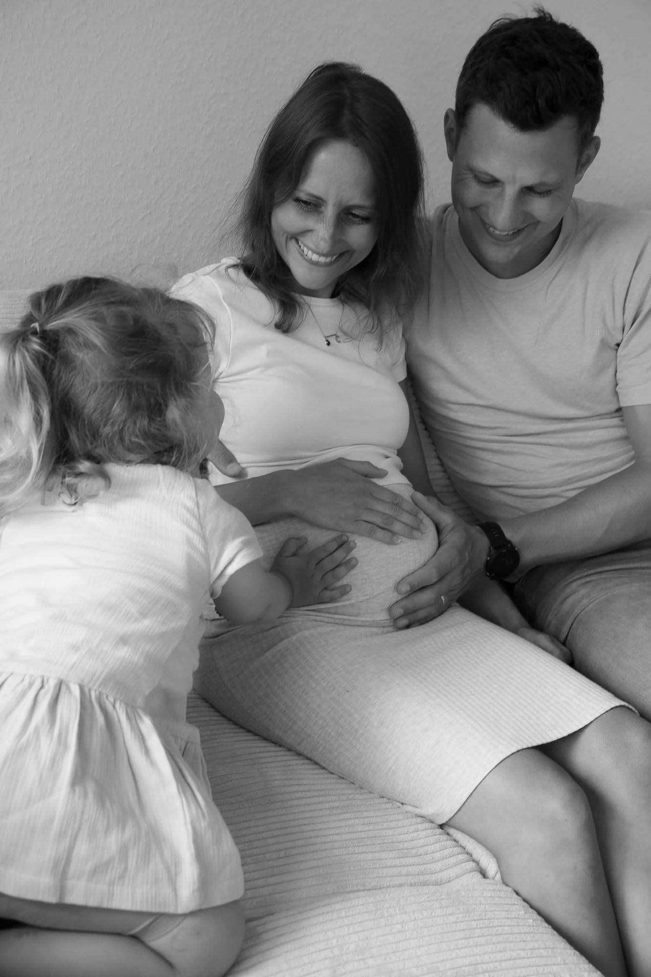 Eine Familie, die ein Baby im Bauch der Mutter spürt, alle lachen und berühren den Bauch, in einem neutralen Raum