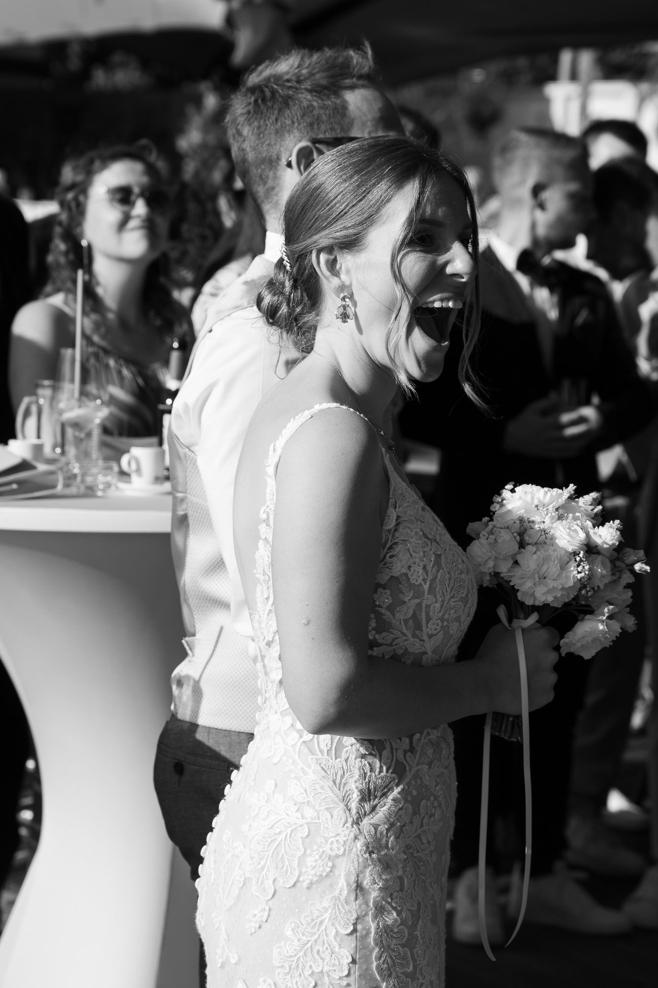 Eine junge Frau in Hochzeitskleid hält einen Blumenstrauß und lacht bei einer Hochzeitsfeier, umgeben von anderen Gästen.