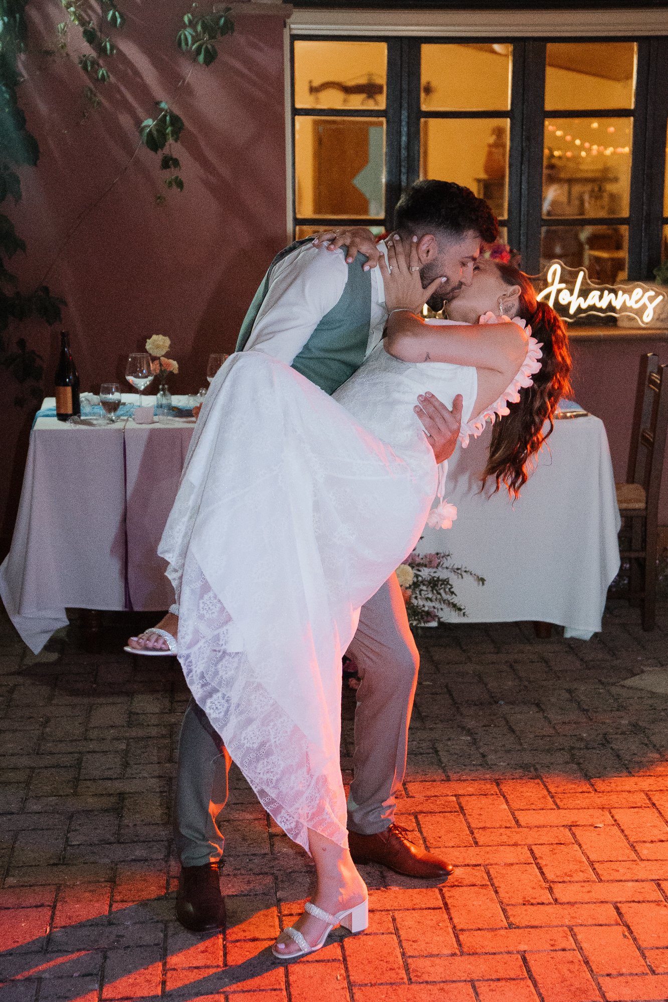 Ein Brautpaar bei einer Hochzeit, der Mann hebt die Frau hoch, beide küssen sich, im Hintergrund ein Tisch mit Getränken und Blumen, eine leuchtende Name-Schriftzug mit 'Johannes' an der Fensterbank, die Szene ist romantisch und festlich.