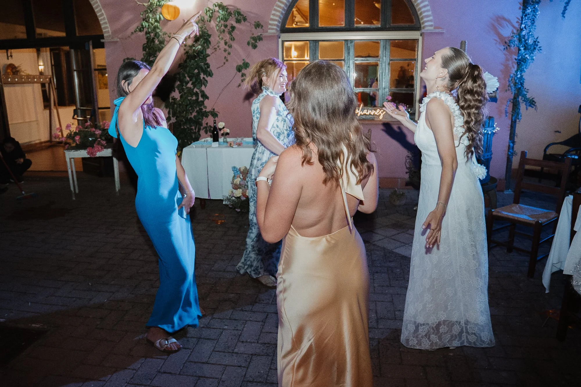Mehrere Frauen tanzen auf einer Party im Freien bei Nacht, mit einem pinkfarbenen Gebäude im Hintergrund.