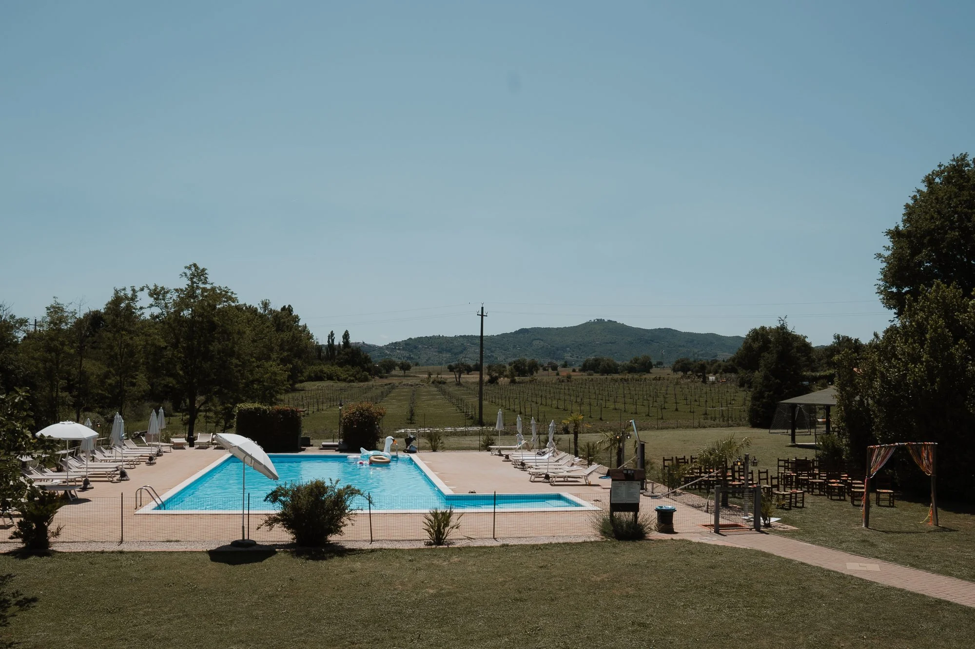 Ein Swimmingpool im Freien mit Sonnenliegen und Sonnenschirmen, umgeben von Bäumen und einer offenen Landschaft mit Hügeln im Hintergrund