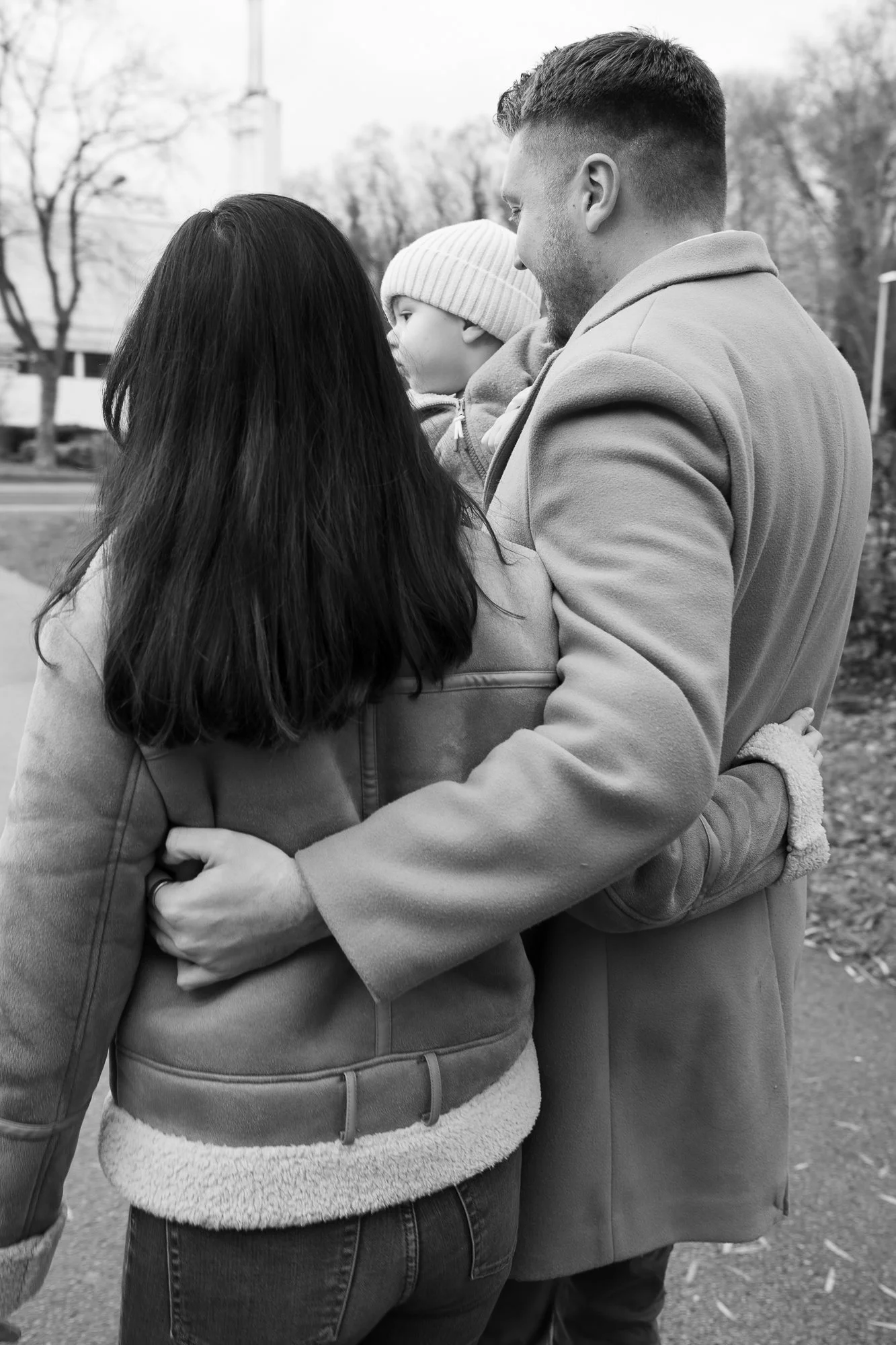Ein Familienbild mit einem Mann, einer Frau und einem kleinen Kind, die sich umarmen, im Freien in einer urbanen Umgebung, in Schwarz-Weiß.