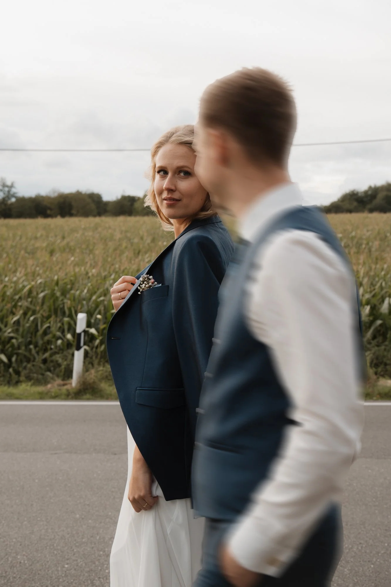 Eine Frau und ein Mann gehen auf einer Landstraße, die Frau schaut zur Kamera, der Mann ist unscharf im Vordergrund. Die Frau trägt ein weißes Kleid und eine dunkle Jacke, der Mann trägt ein weißes Hemd und eine dunkle Weste.