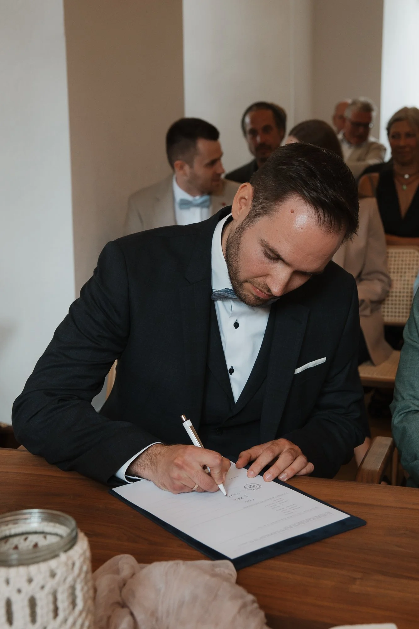 Mann in Schwarzem Anzug und Fliege schreibt eine Urkunde bei einer offiziellen Zeremonie, während im Hintergrund mehrere Menschen zuschauen.