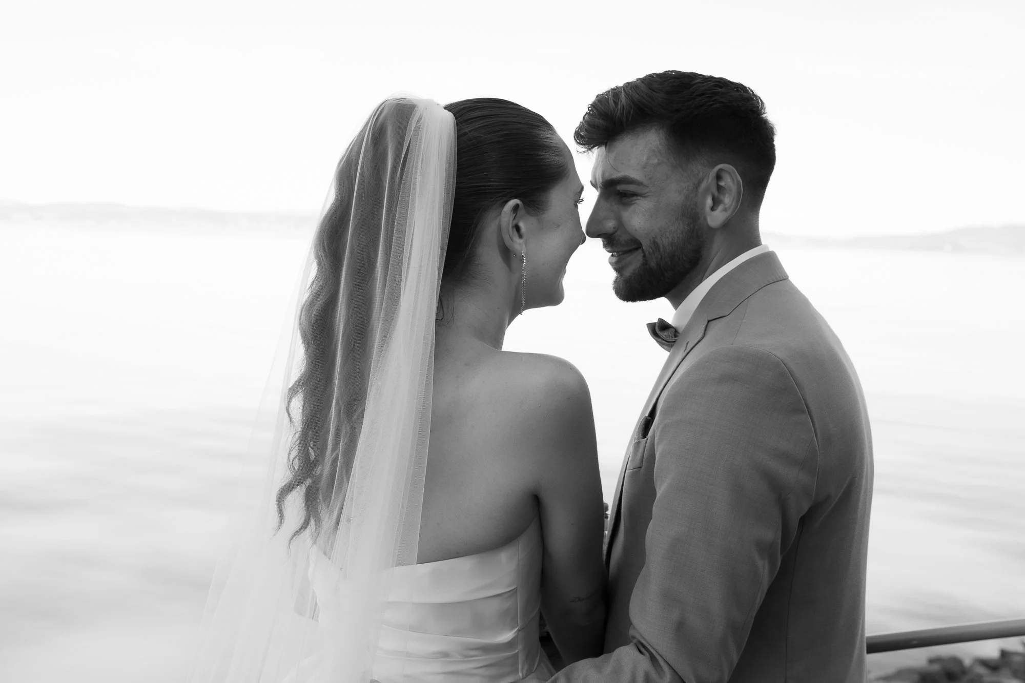 Ein Brautpaar steht eng beieinander am Wasser, lacht und blickt sich in die Augen. Die Braut trägt ein wehendes Tüllkleid und einen Schleier, der über ihre Haare fällt. Der Bräutigam trägt einen Anzug mit Fliege.