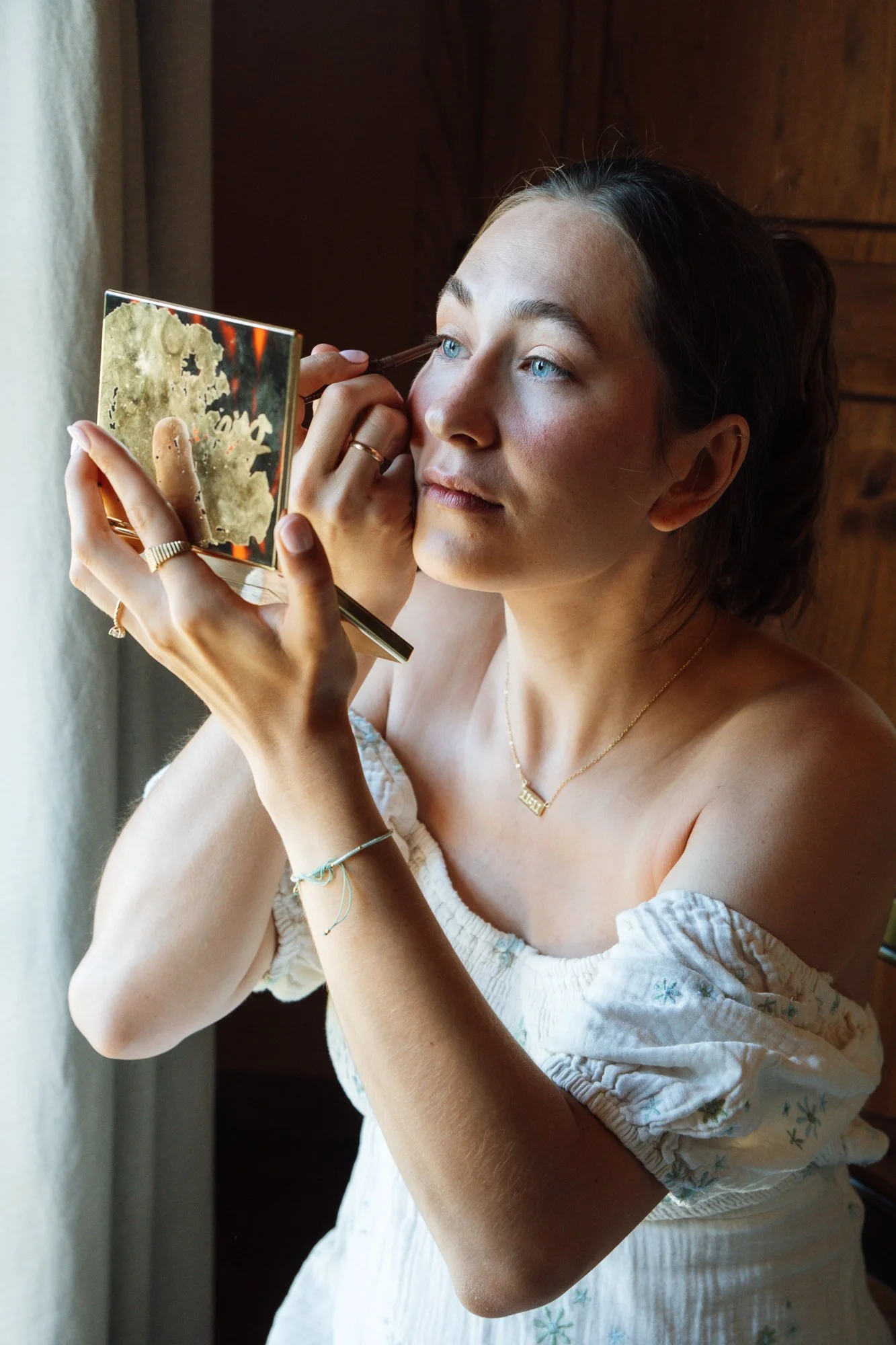 Junge Frau beim Make-up, die vor einem Fenster sitzt und sich im Spiegel betrachtet.