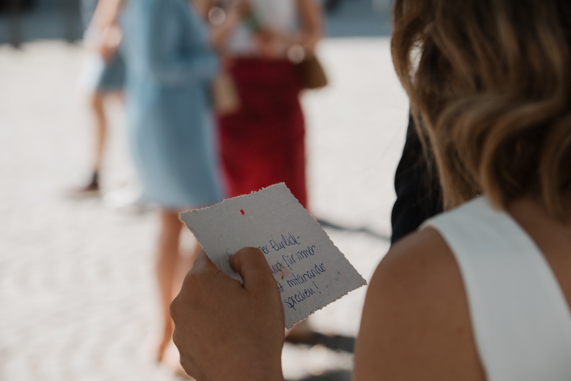 Frau liest einen handgeschriebenen Zettel auf einem Strand, im Hintergrund sind andere Menschen in Sommerkleidung, die im Sand stehen.