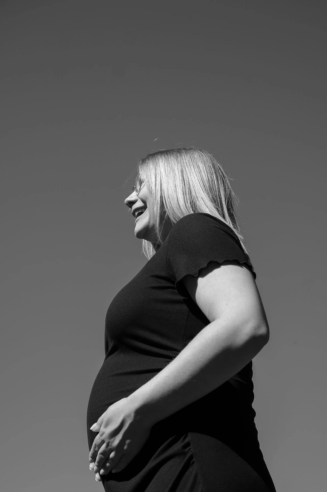 Eine schwangere Frau lacht und schaut nach oben auf einem schwarzen Kleid, im Freien bei klarem Himmel.