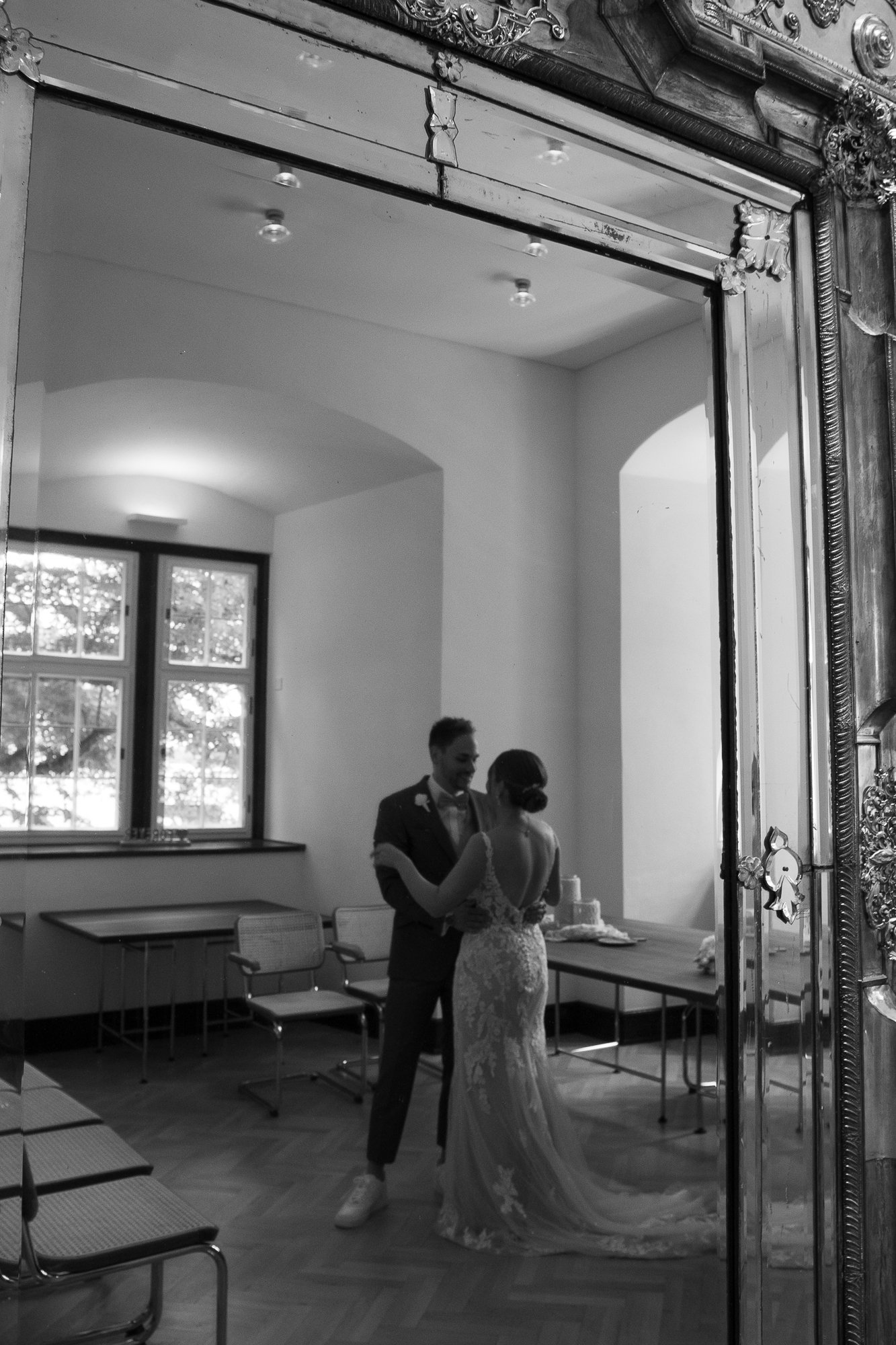 Ein Brautpaar tanzt im Spiegel, im Hintergrund eine Fensterfront mit Bäumen, auf einem Tisch eine Hochzeitstorte.