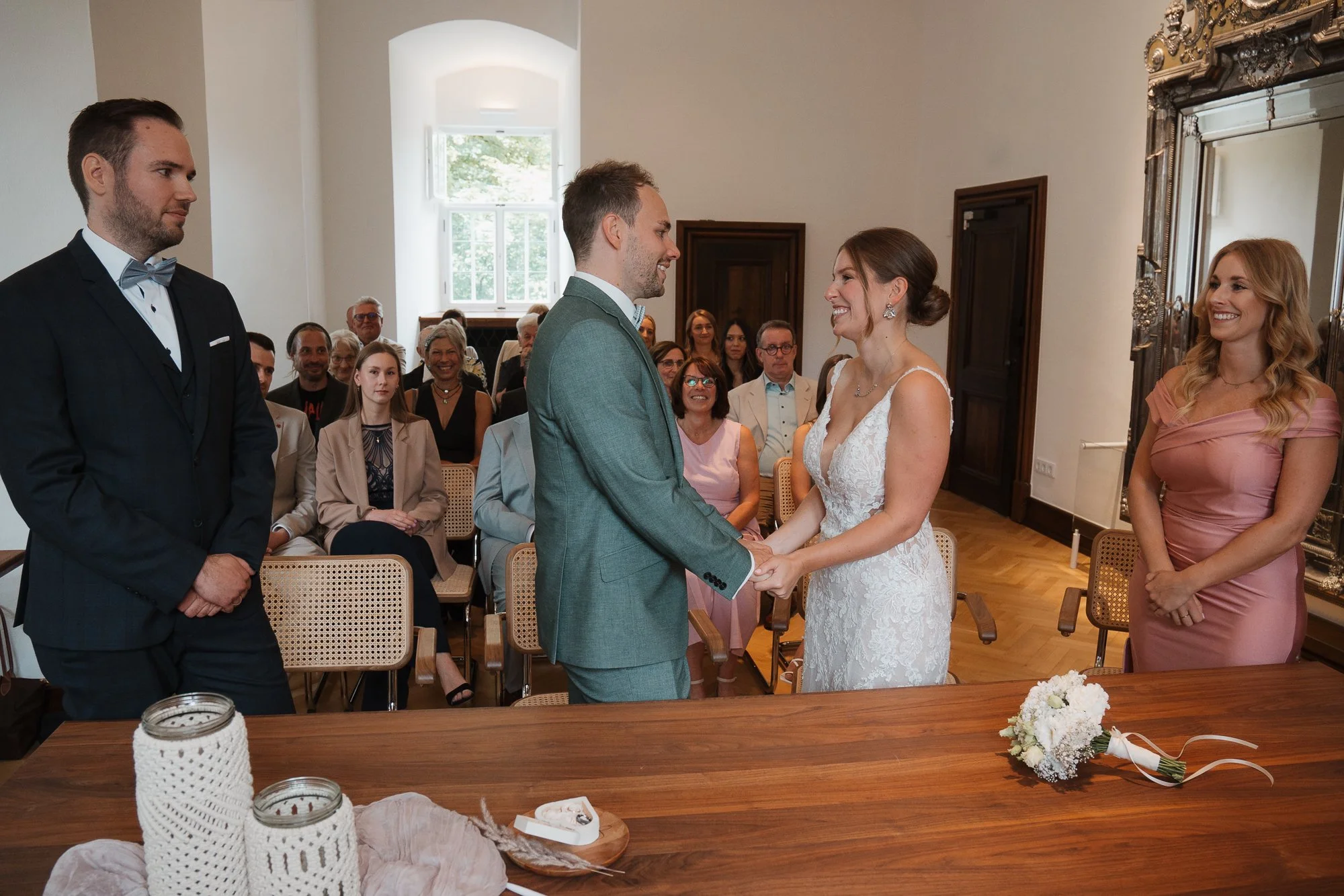 Ein Brautpaar bei der Trauung, das sich gegenseitig die Hände hält, in einem festlichen Raum mit Gästen im Hintergrund.