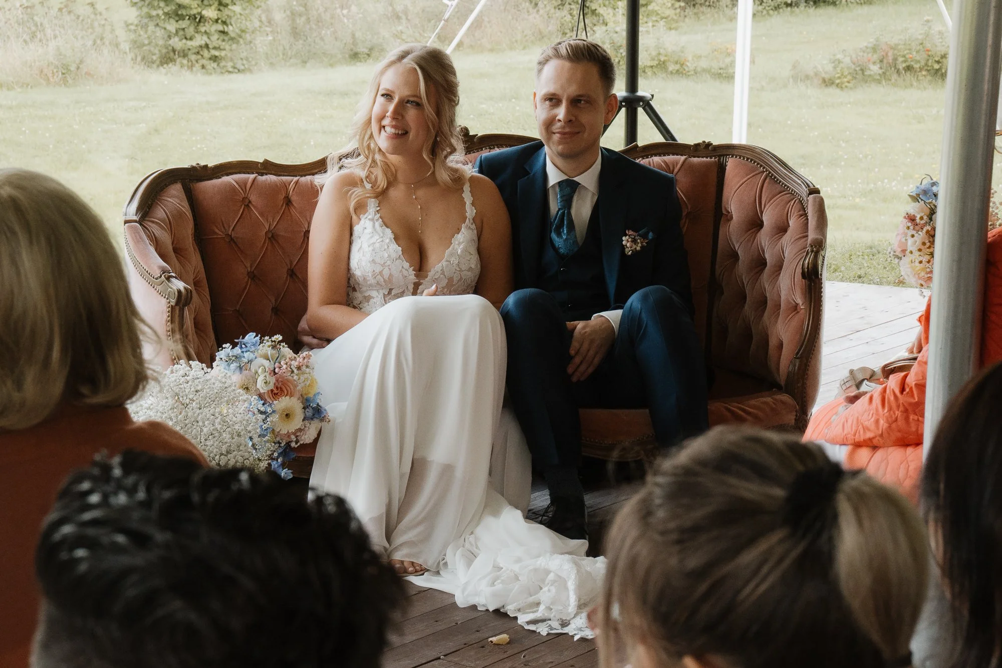 Braut und Bräutigam sitzen auf einem roten Samtsofa bei einer Hochzeit, umgeben von Gästen, im Hintergrund eine grüne Wiese im Freien.