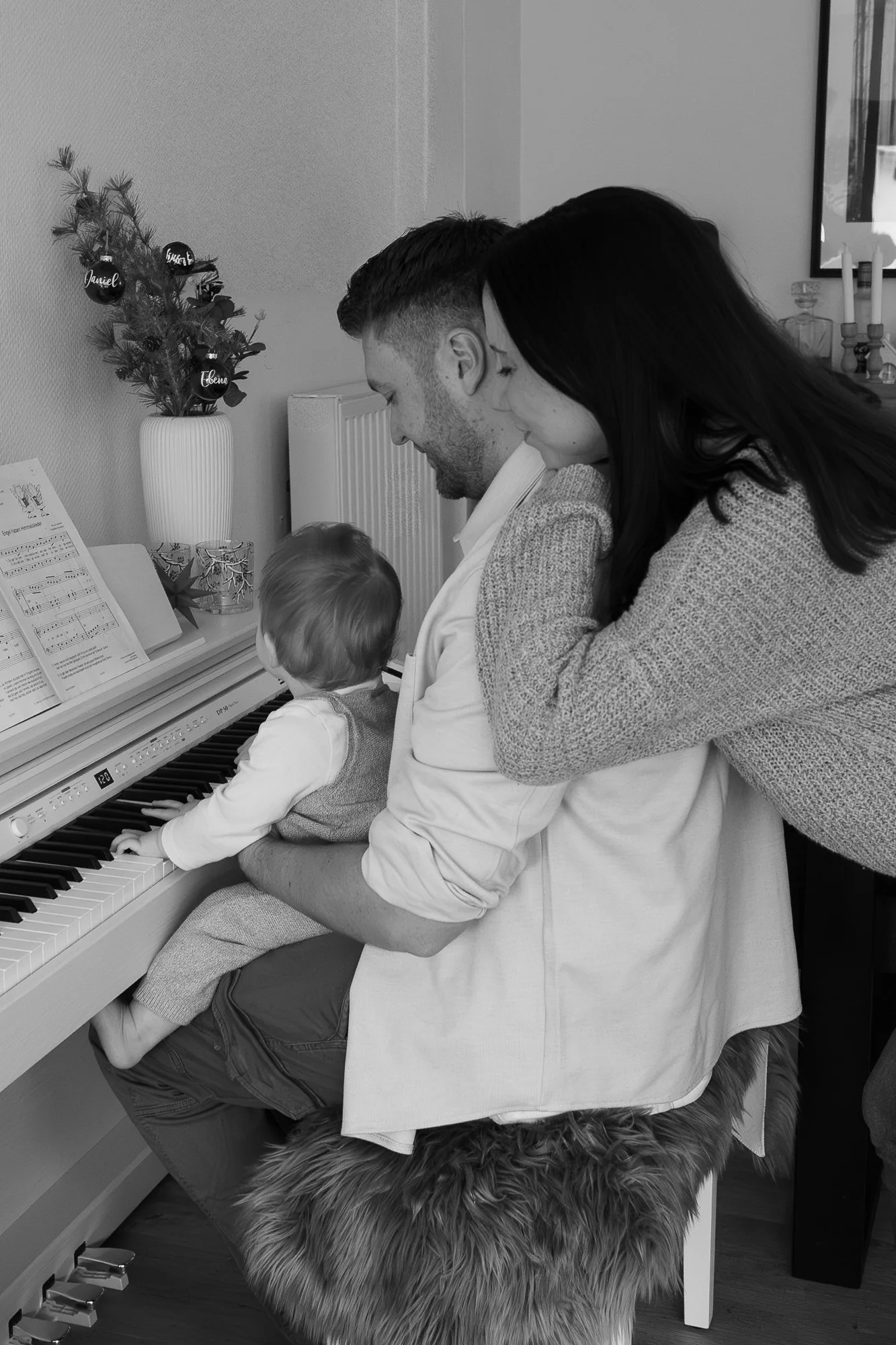 Eine Familie mit einem kleinen Kind, das Klavier spielt, während die Eltern Zuschauen, in einem gemütlichen Wohnzimmer mit Weihnachtsdekoration im Hintergrund.