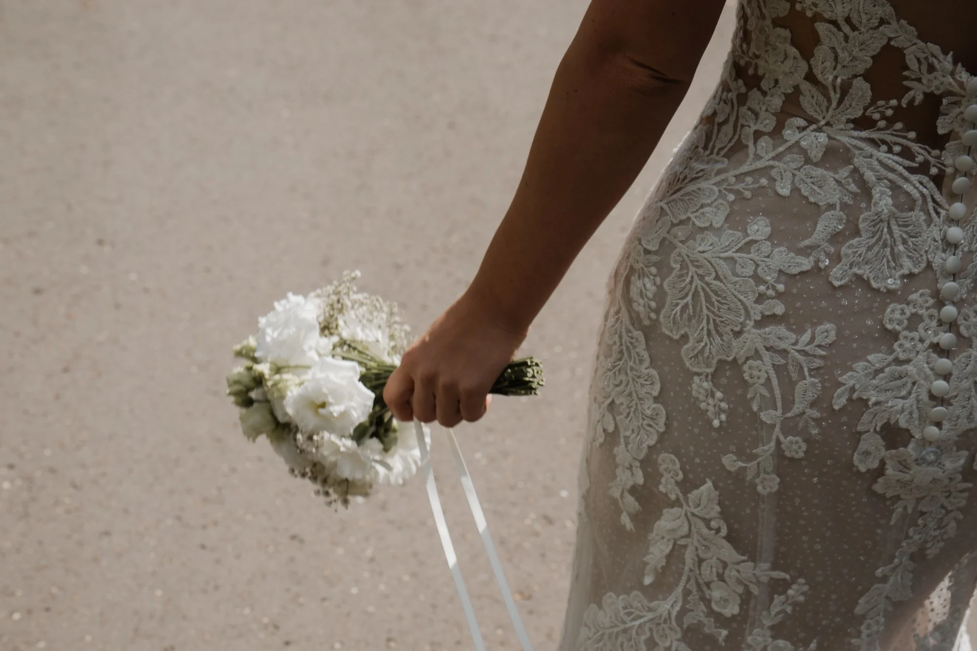 Eine Person trägt ein hochzeitskleid, das mit Spitzenverzierungen und Knöpfen auf der Rückseite versehen ist, während sie ein weißes Brautstrauß hält.