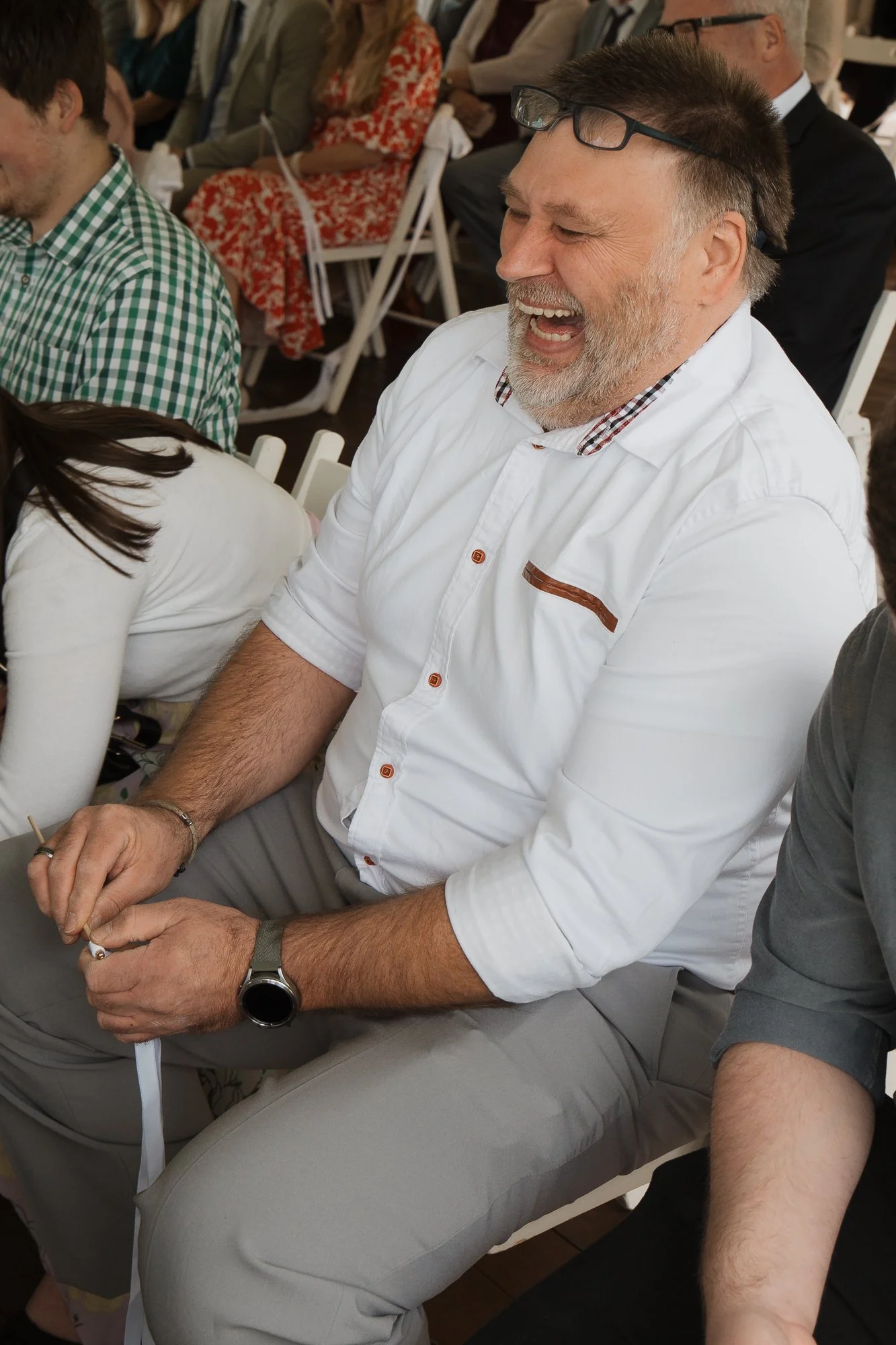 Ein lachender Mann mit Bart und Brille auf dem Kopf sitzt an einer Veranstaltung, umgeben von anderen Menschen.