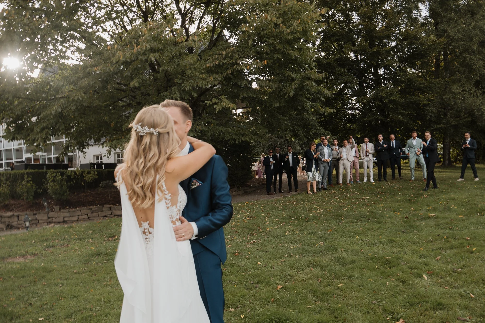 Ein Hochzeitsfoto, auf dem ein Brautpaar sich küsst, während die Gäste im Hintergrund bei einer Feier im Freien stehen, umgeben von Bäumen.