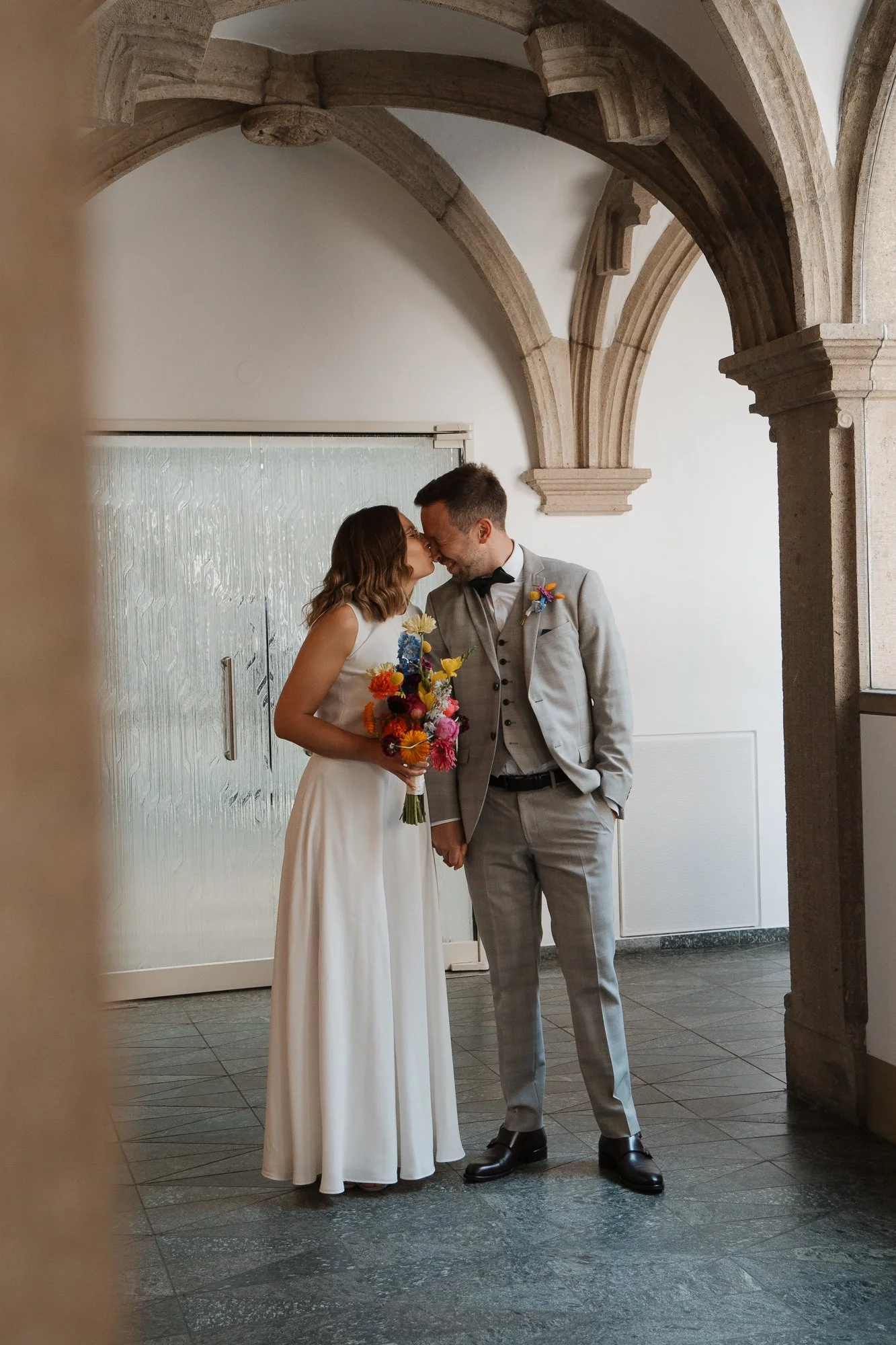 Ein heiratswilliges Paar bei einer Zeremonie, die sich küssen, im Hintergrund eine helle Wand und einen steinernen Bogen, sie halten eine bunte Blumenstrauß