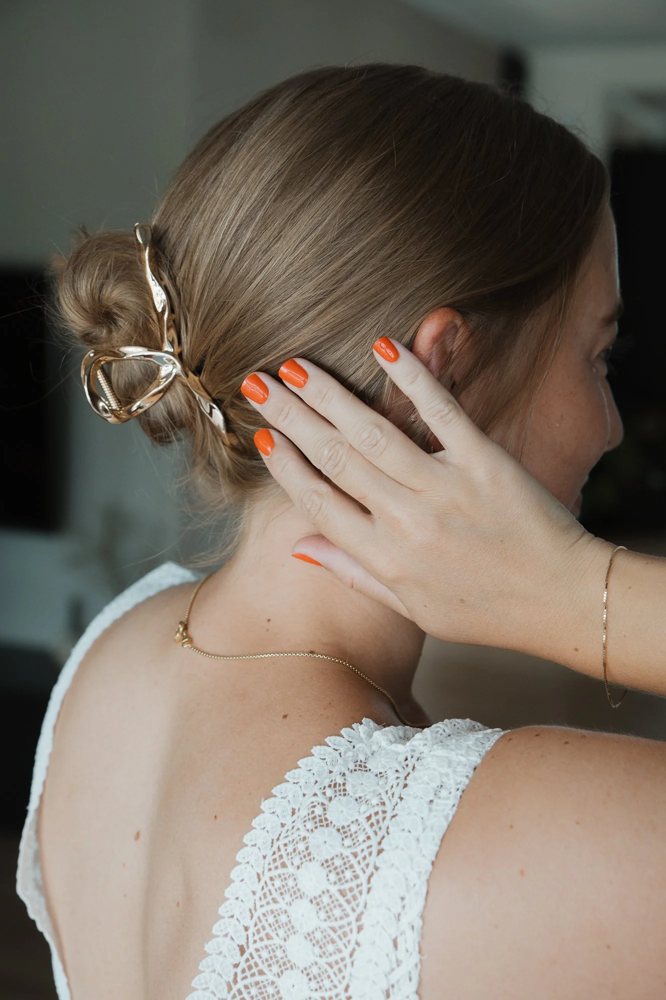 Nahaufnahme einer Frau, die ihre Haare mit einer großen Haarklammer stylt, trägt eine weiße Spitzebluse, und hat rote Nagellacke. Im Hintergrund ist ein unscharfer Raum zu sehen.