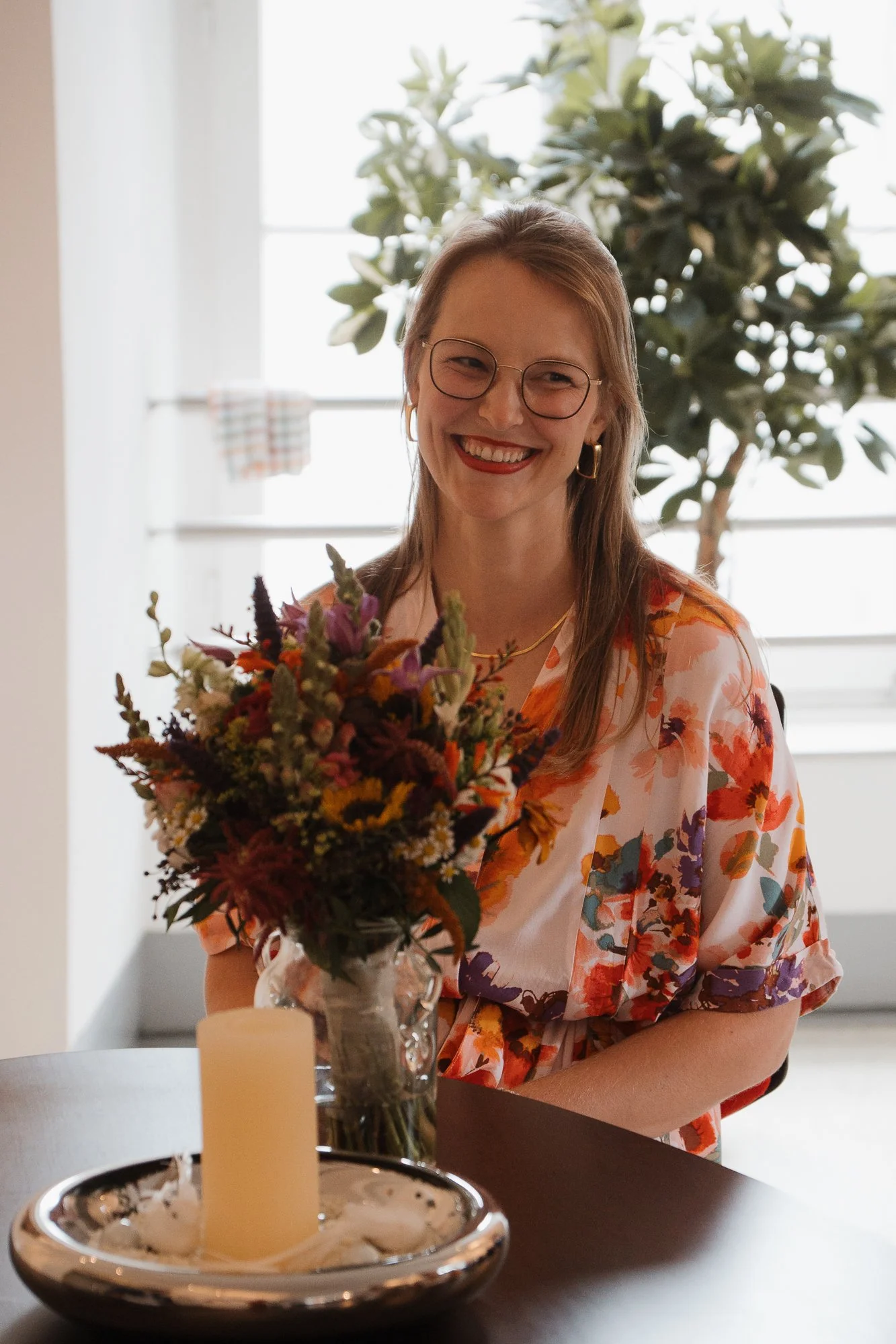 Eine lachende Frau mit Brille, Ohrringen und einem bunten, floralen Kleid sitzt an einem Tisch mit einem Blumenstrauß und einer Kerze.