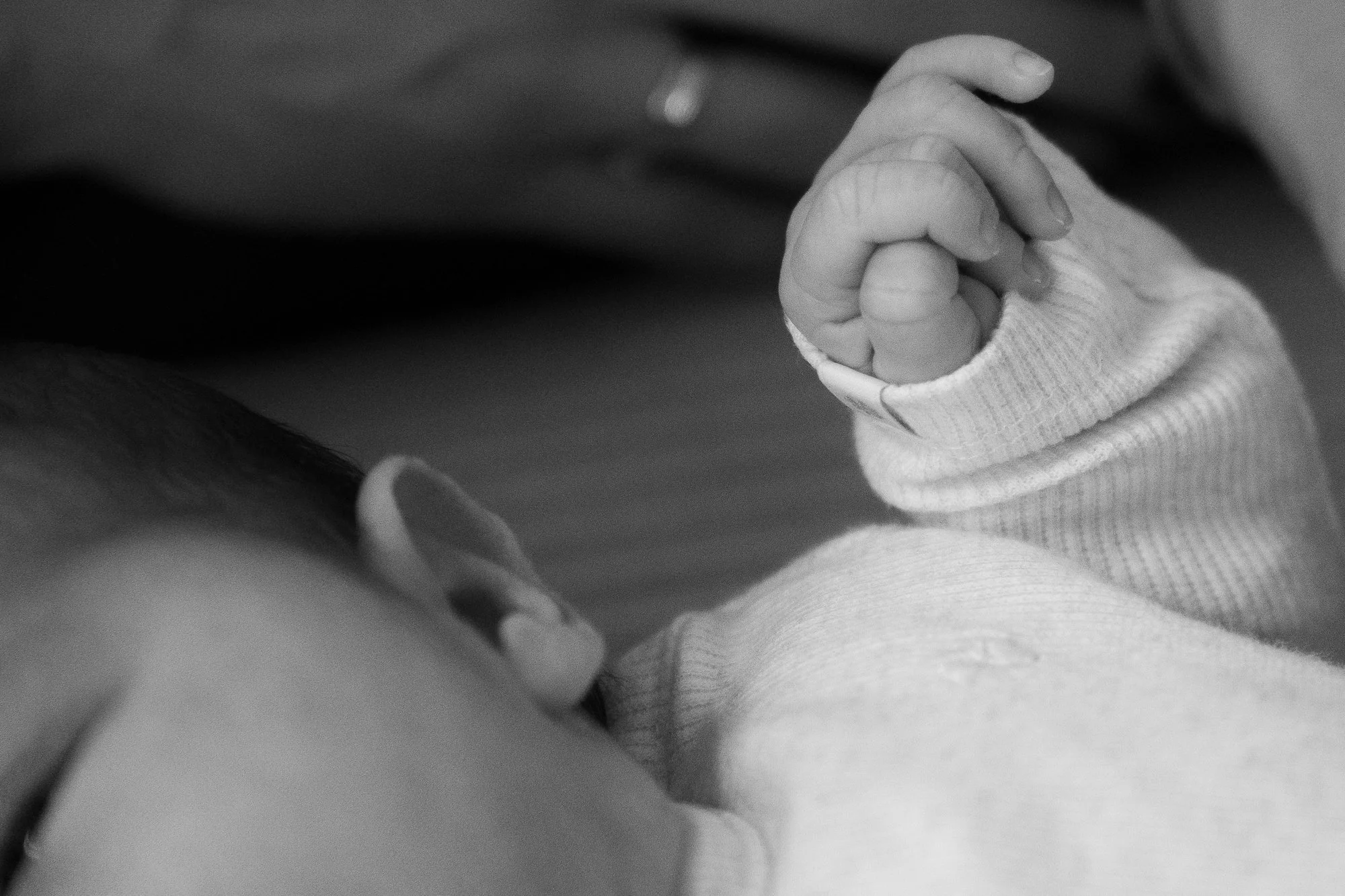 Nahaufnahme einer Hand eines Neugeborenen mit Daumen nach oben, auf einem Arm eines Erwachsenen liegend, in Schwarz-Weiß.