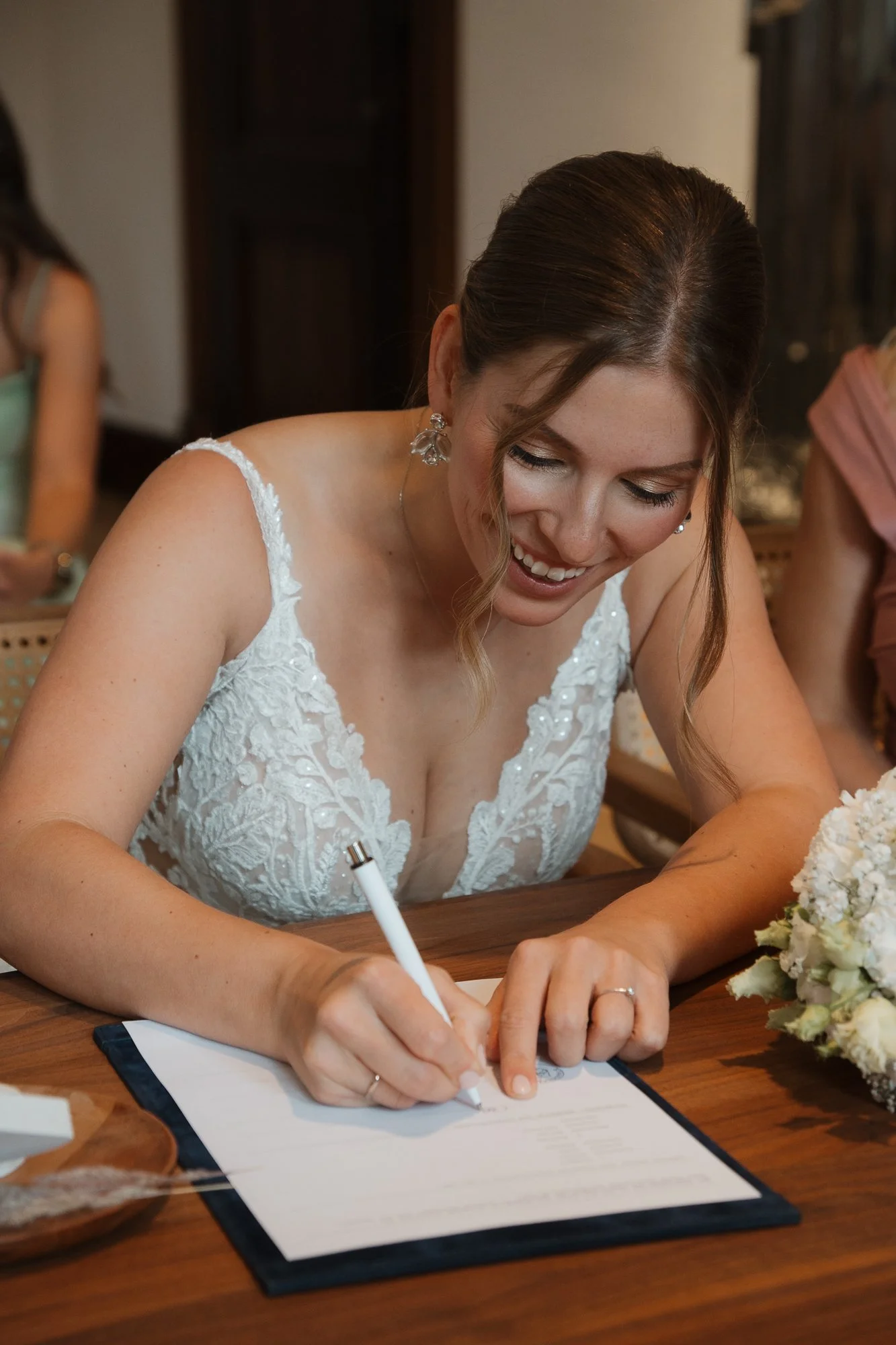 Eine Braut unterschreibt ein Dokument an einem Tisch, trägt ein weißes Hochzeitskleid, hat braune Haare und trägt Schmuck.