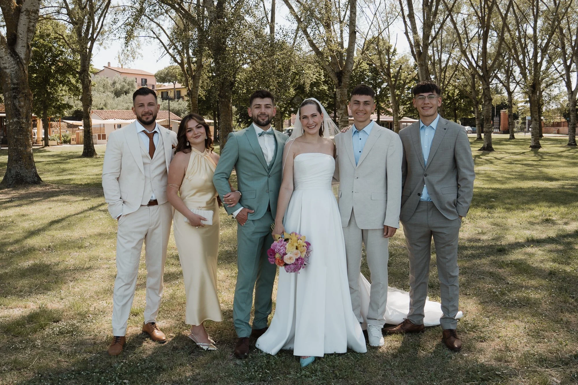 Eine Gruppe von sieben Menschen, die bei einer Hochzeit im Freien stehen, darunter die Braut in Weiß mit Blumenstrauß und die Gäste in eleganten Anzügen und Kleidern.