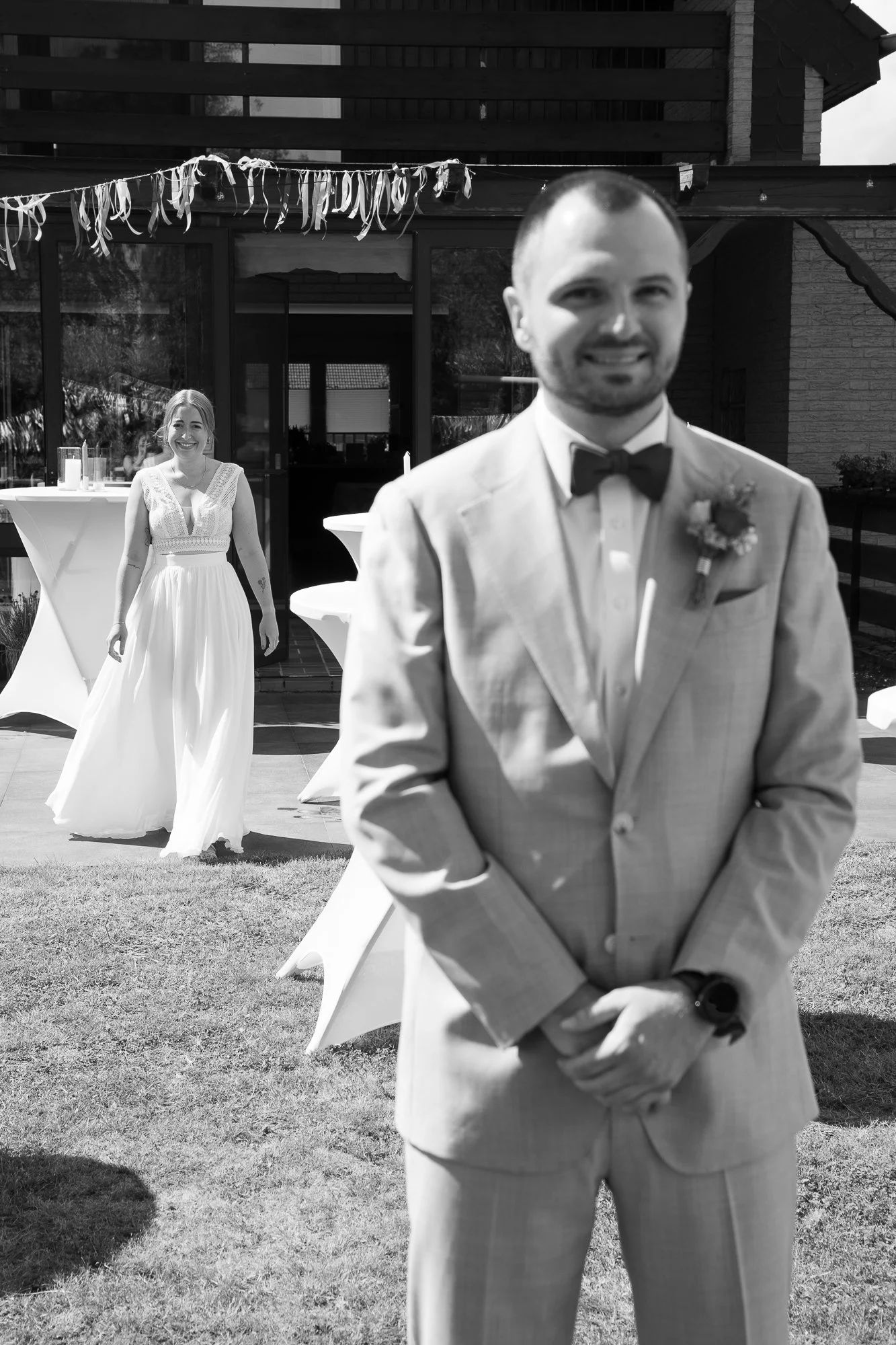 Ein Mann in einem Anzug mit Fliege und einer Frau in einem weißen Kleid bei einer Hochzeitsfeier im Freien, im Hintergrund ein Haus.